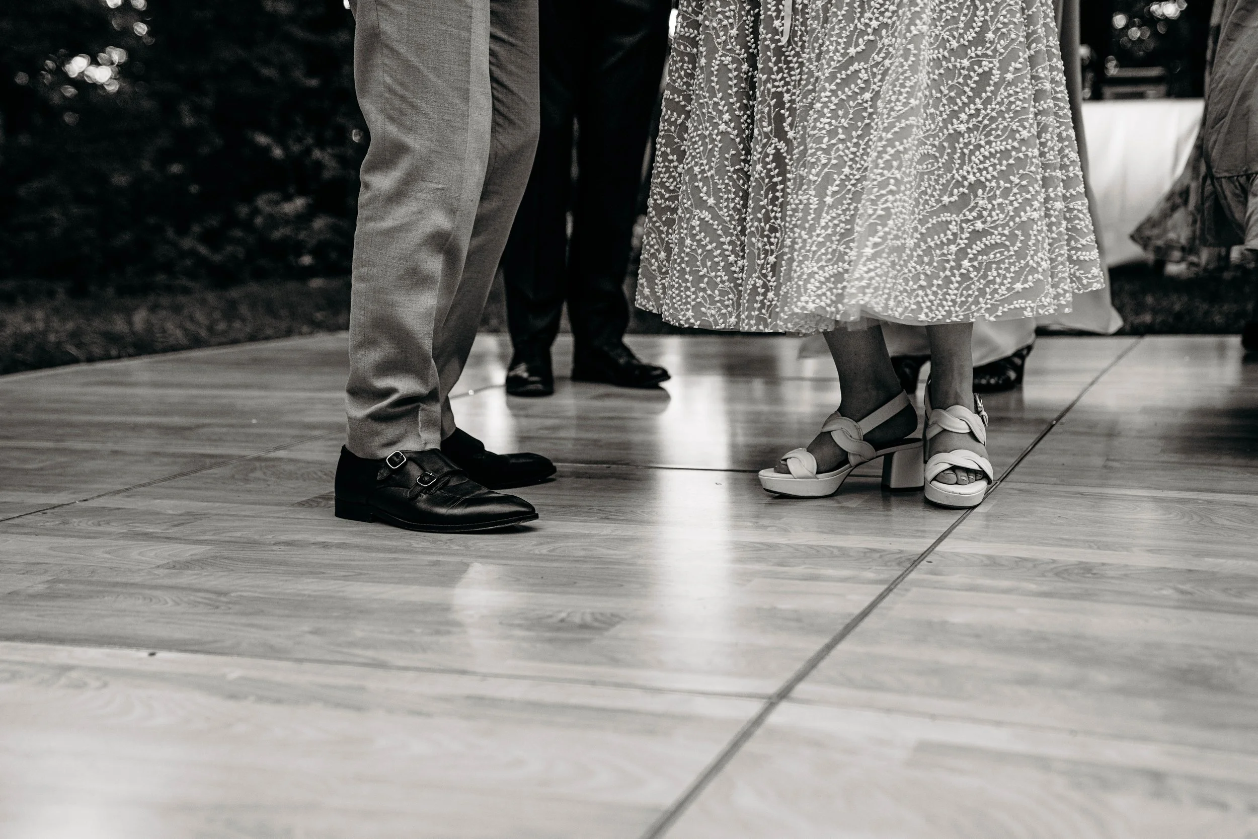 Close-up of people's feet, including a woman wearing high-heeled sandals and a man in formal shoes, standing on a dance floor.
