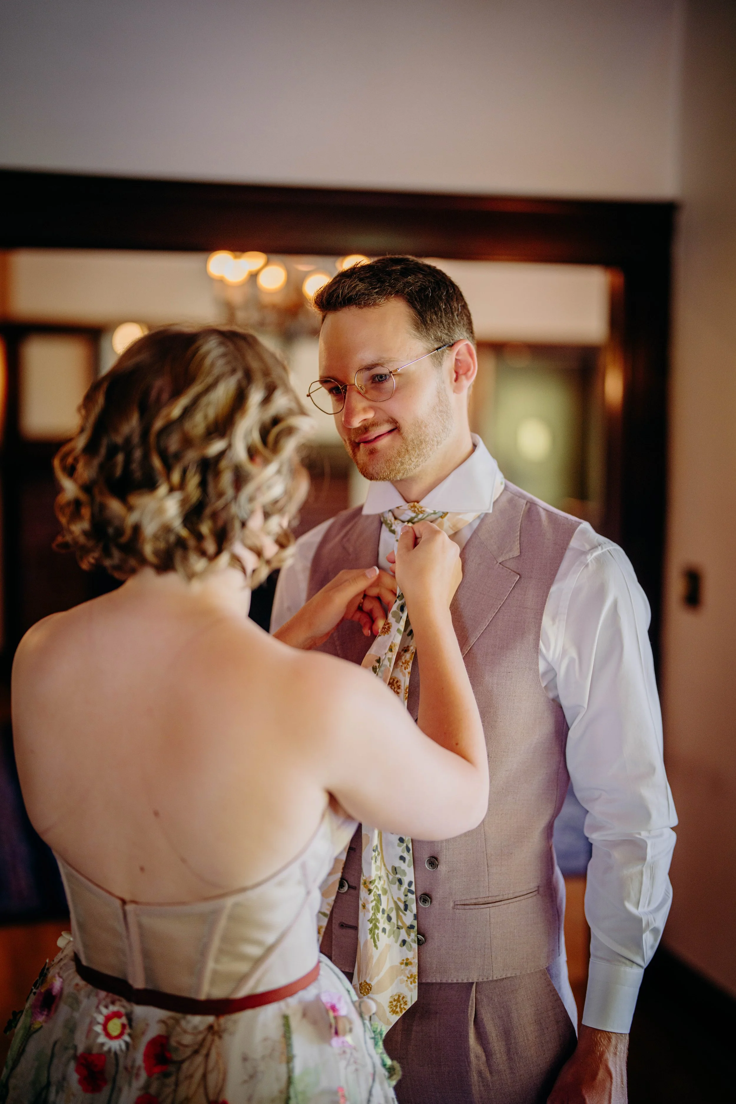 A woman with curly hair is helping a man tie his necktie. The man is wearing glasses and a light purple vest over a white shirt, and the woman has short hair and is wearing a strapless dress with floral patterns.