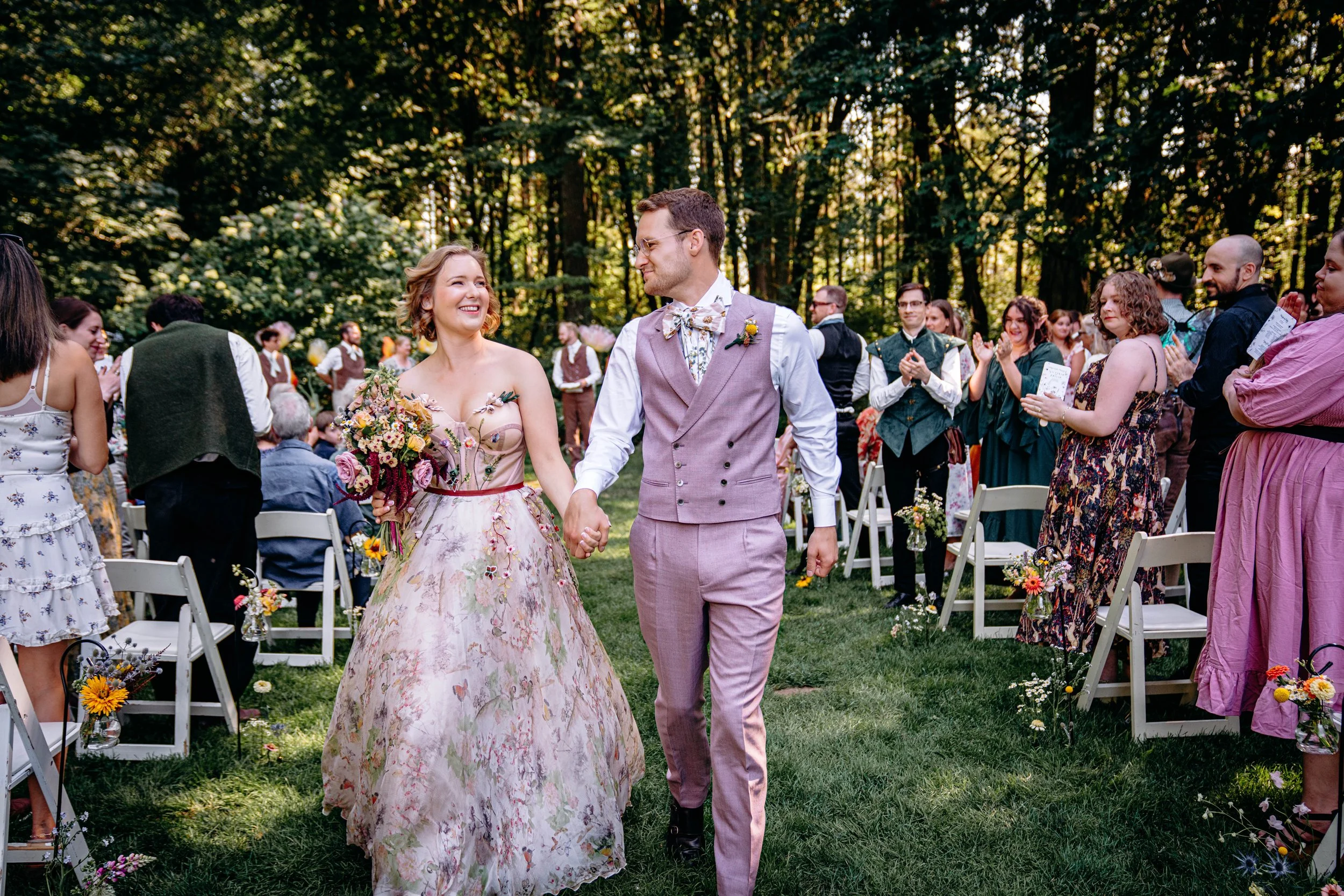 A bride and groom walking hand in hand down a grassy aisle at an outdoor wedding ceremony, surrounded by applause and smiles from the guests, set in a lush green forest.