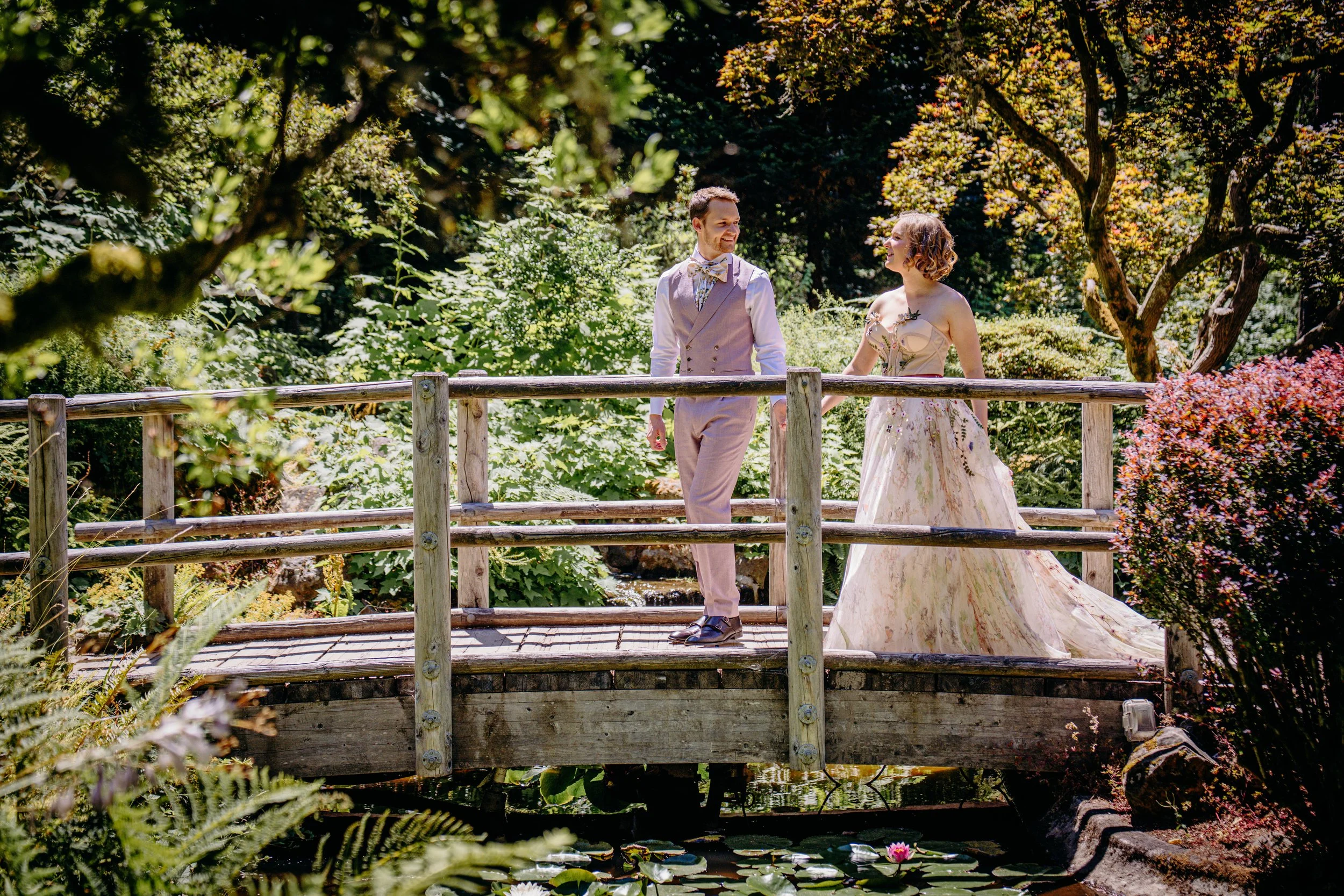 A man and woman holding hands on a wooden bridge in a lush garden, dressed in vintage wedding attire, smiling at each other.
