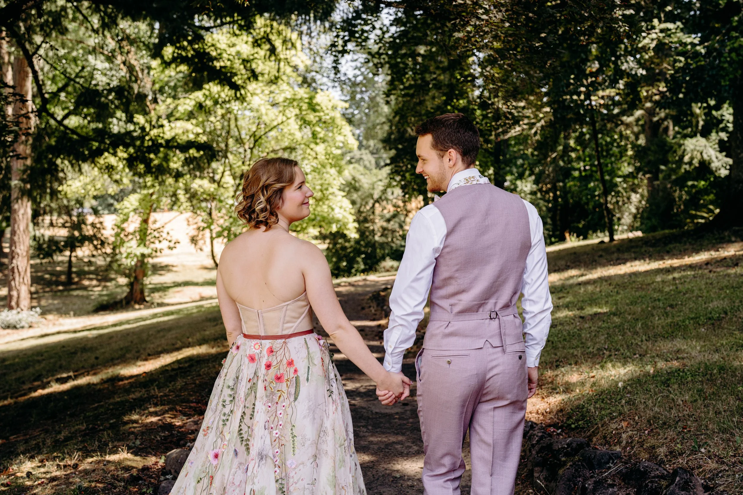A couple holding hands in a forest during daytime, with the woman in a strapless floral dress and the man in a light purple vest and pants, both smiling at each other.