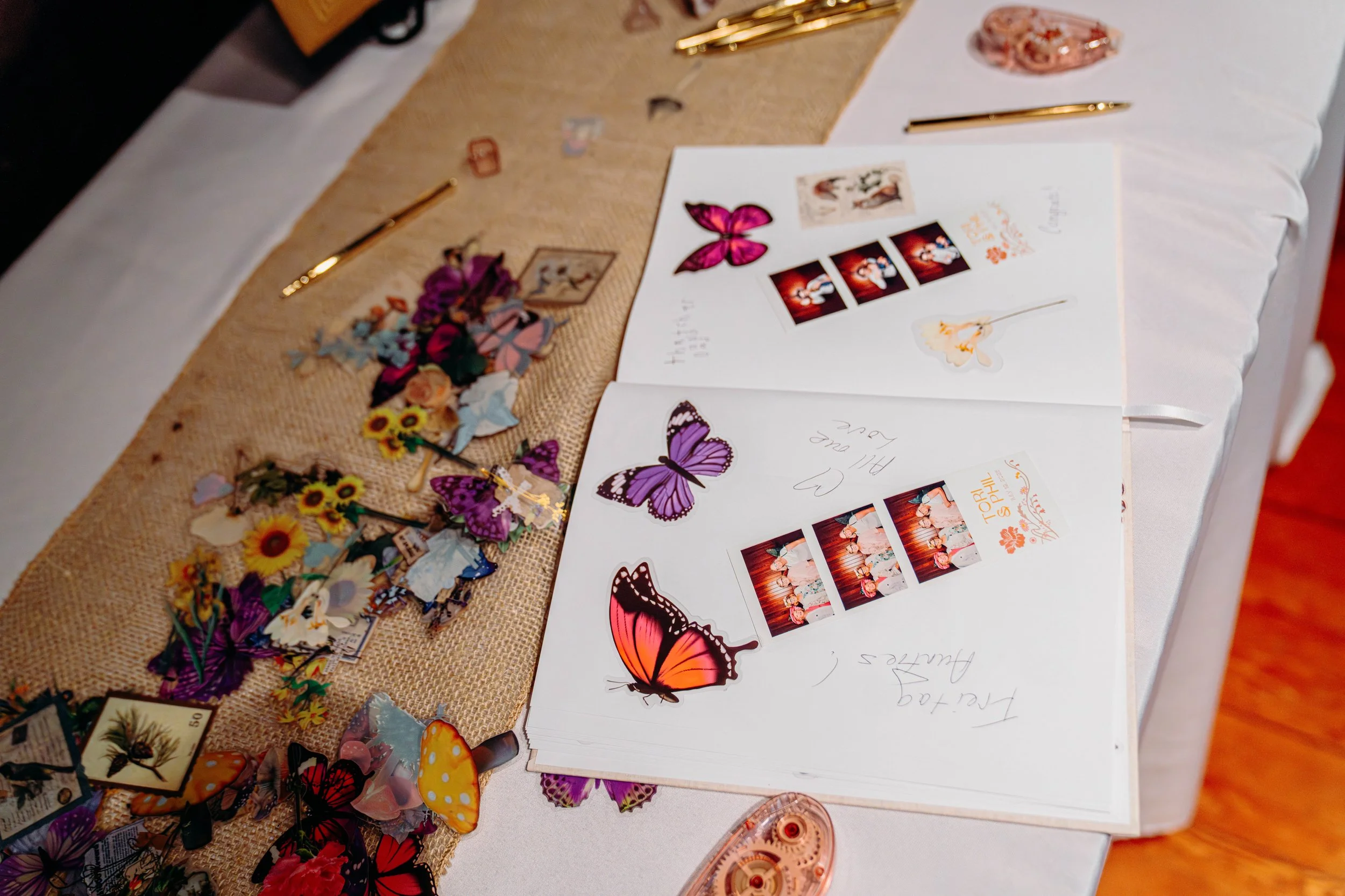 Open photo album on a table with butterfly stickers and photo booth strips. The table has a burlap runner and some flowers. There are pens and a decorative pin on the table.