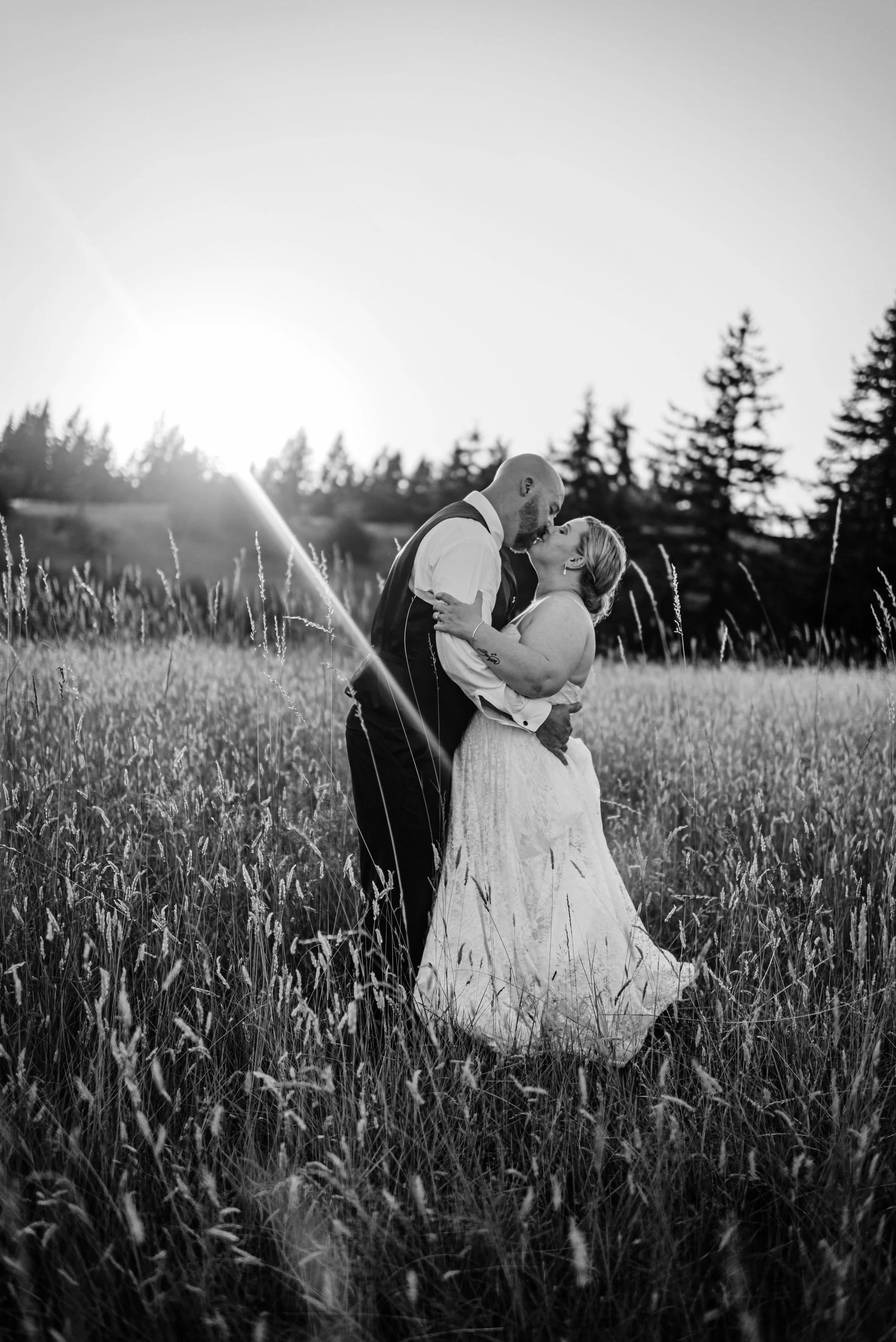 A black and white photo of a couple kissing in a field at sunset, with trees in the background.