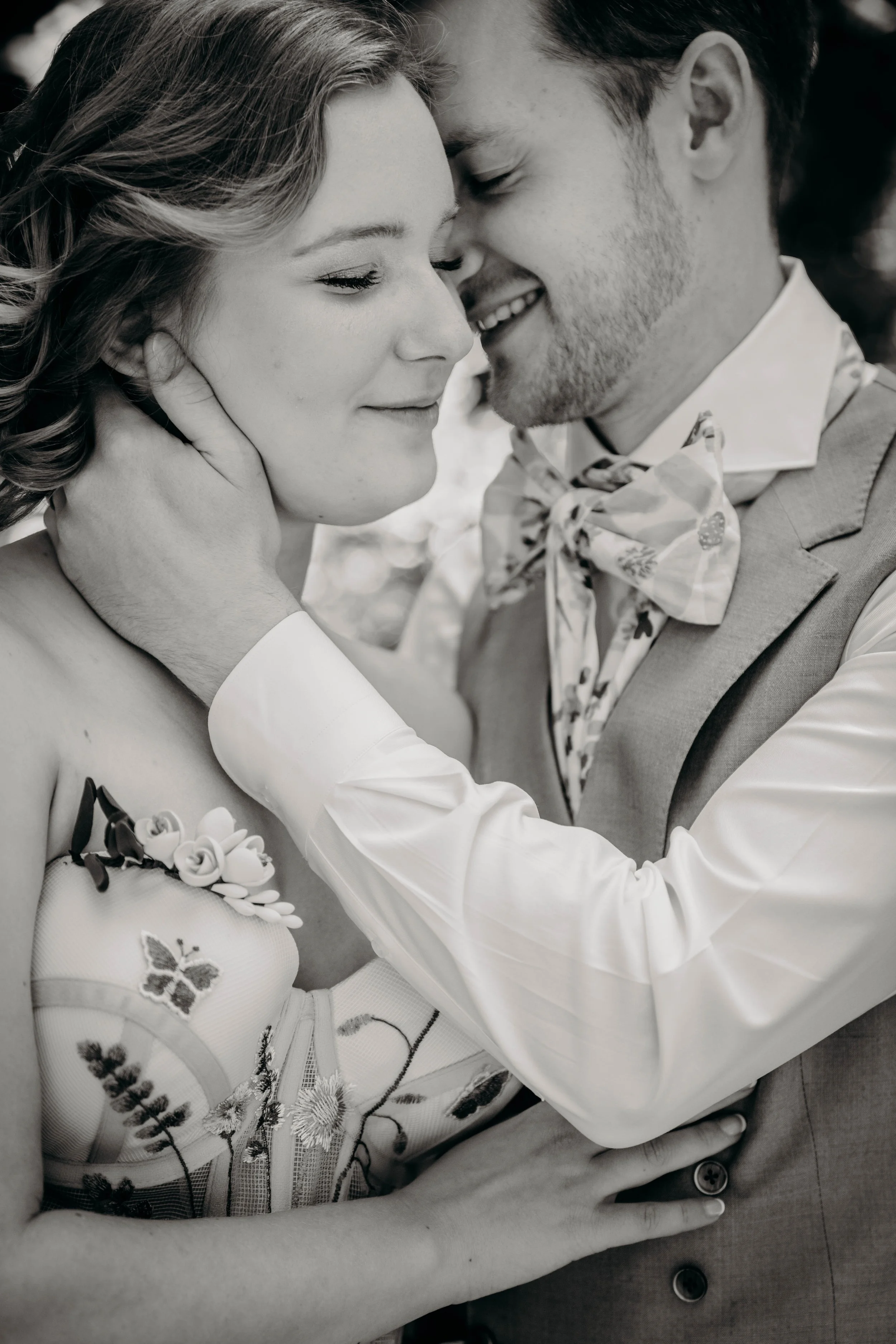 A black and white photo of a couple with eyes closed, noses touching, and smiling warmly. The woman has light-colored hair and is wearing a floral dress with embroidered details, and the man is in a suit with a patterned bow tie. They are embracing c