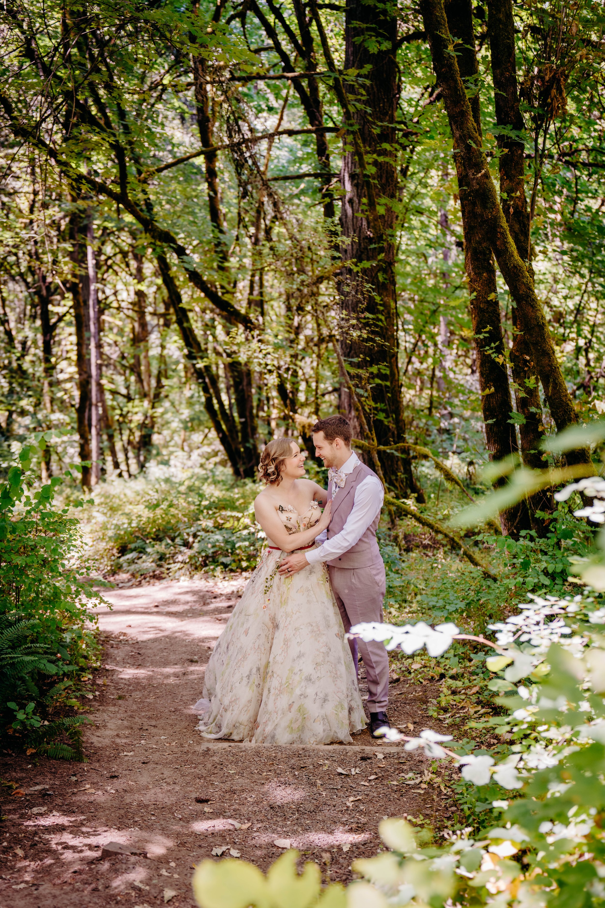 A couple in wedding attire standing together on a forest trail surrounded by lush green trees and foliage, smiling at each other.