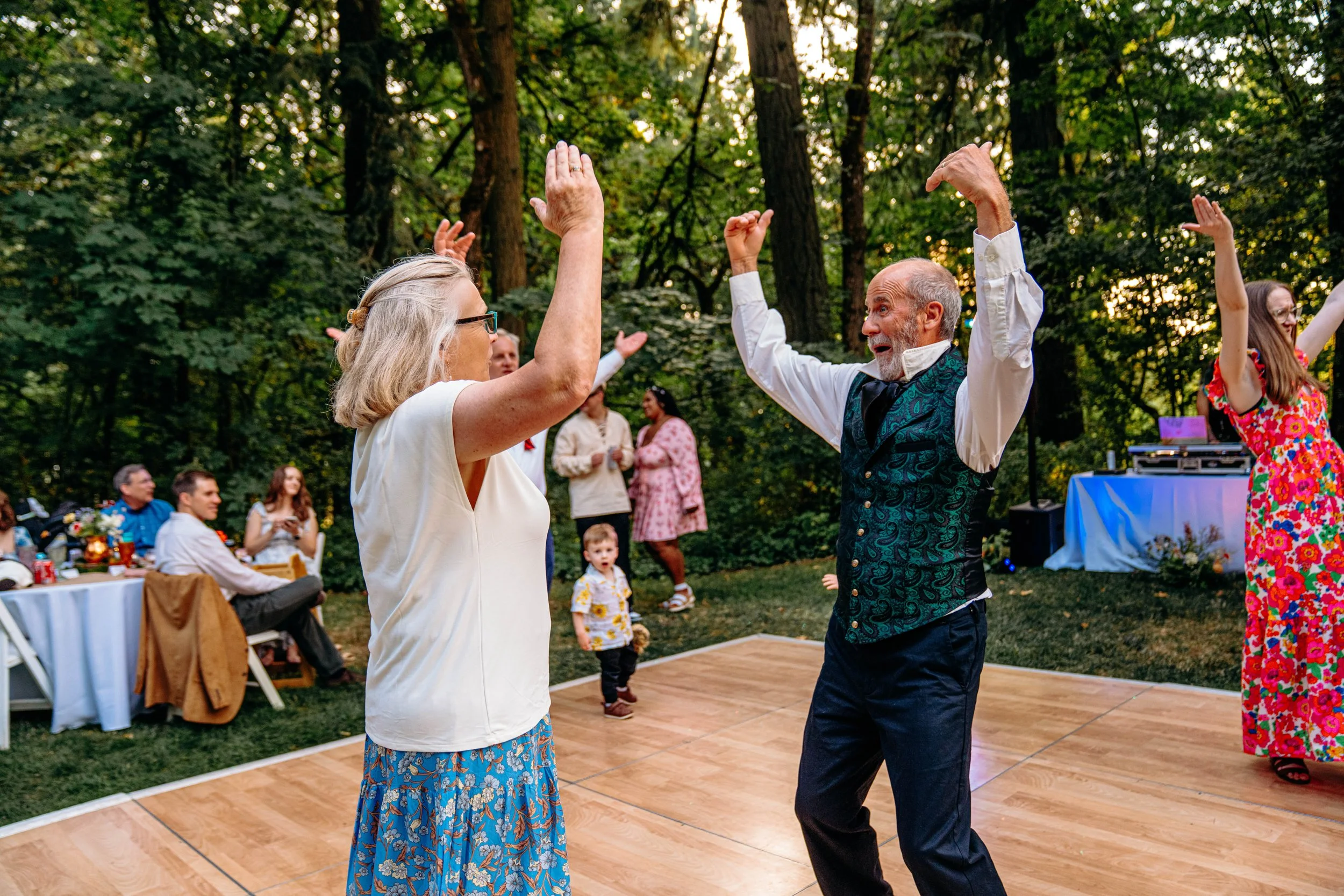 Older couple dancing on a wooden dance floor outdoors at a garden party. The woman is wearing a white top and a blue patterned skirt, and the man is in a white shirt with a dark vest. Other guests sit and stand around, some watching and chatting, wit