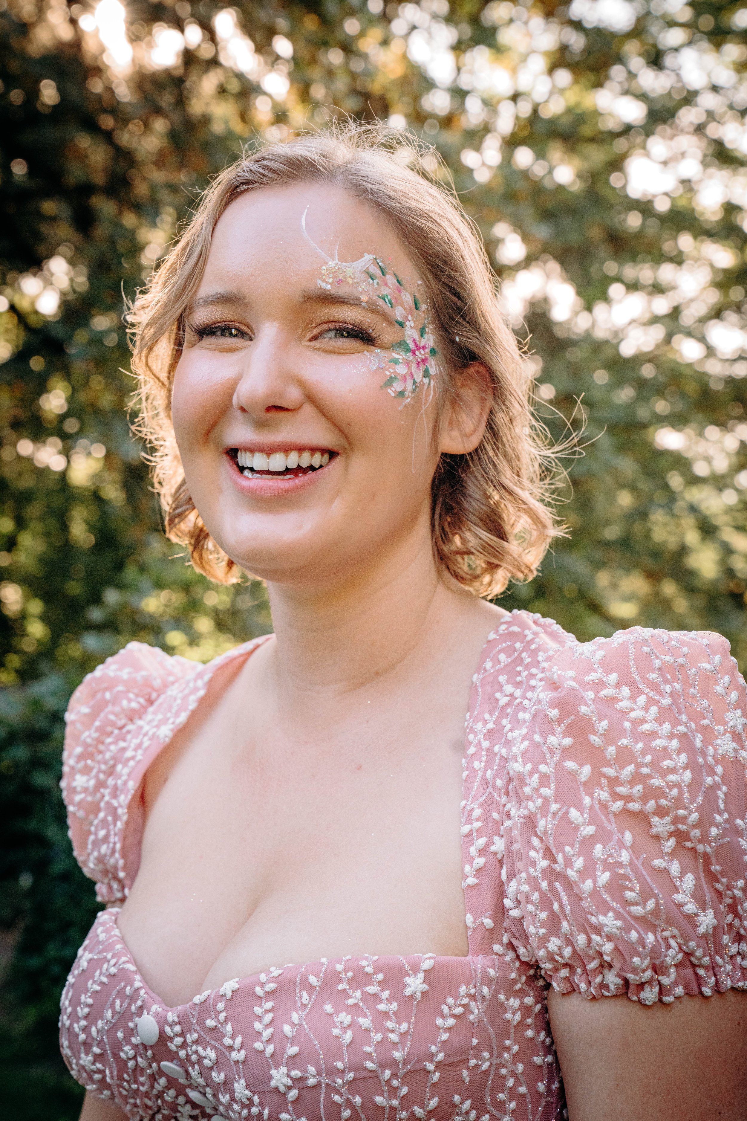 A young woman with short, wavy light brown hair smiling outdoors, with floral face painting on her left temple and wearing a pink dress with white embroidery, against a background of trees and sunlight.