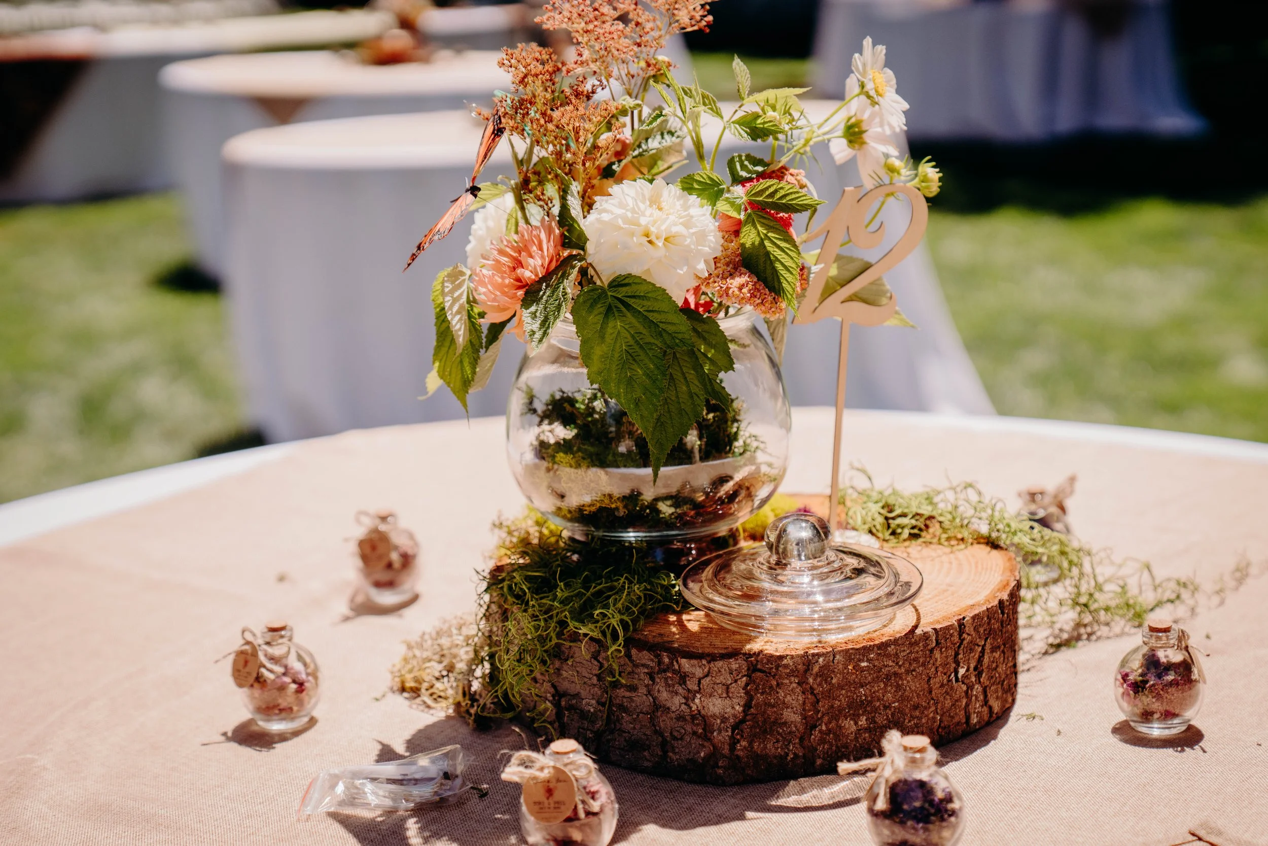 A floral arrangement in a glass bowl sits on a tree trunk slice, decorated with small jars and tags on a round table at an outdoor event.