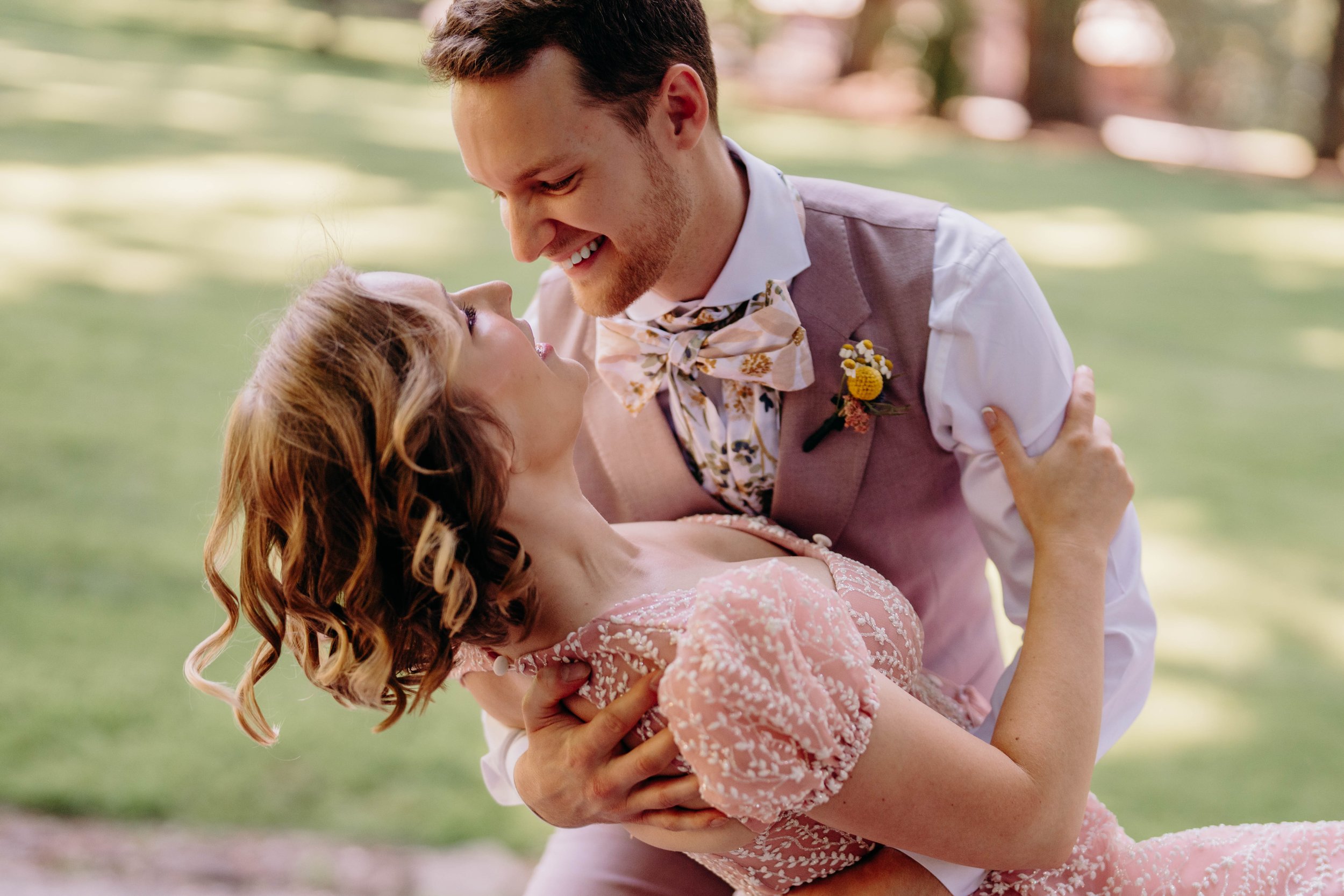 A couple dancing outdoors on a sunny day, dressed in vintage style, with the man in a vest and bow tie, and the woman in a pink dress with ruffled sleeves.
