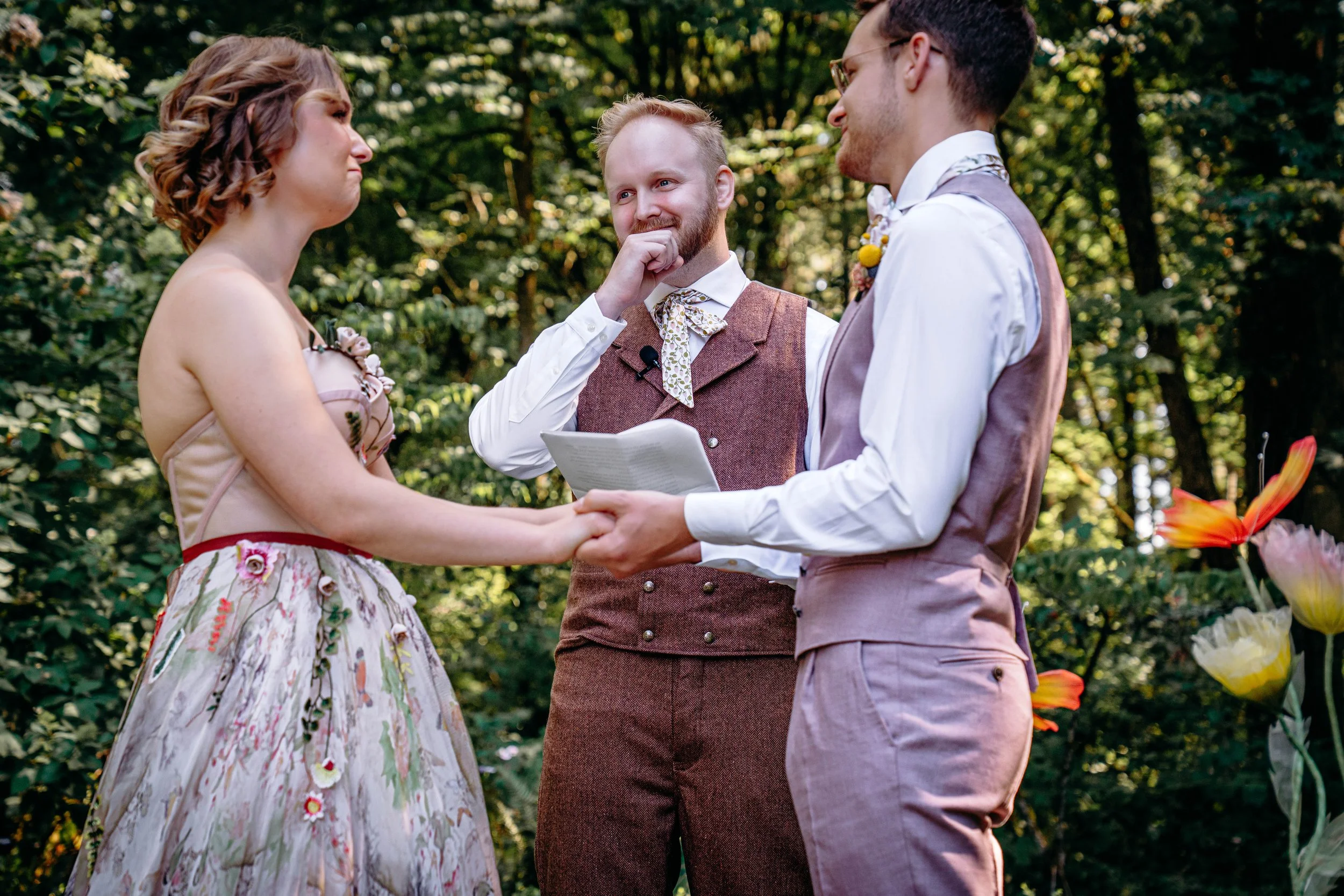 A couple is getting married outdoors, holding hands and facing each other, with a officiant in between them. The bride is wearing a floral dress with floral embellishments, and the groom is dressed in a light pink vest and trousers with a white shirt