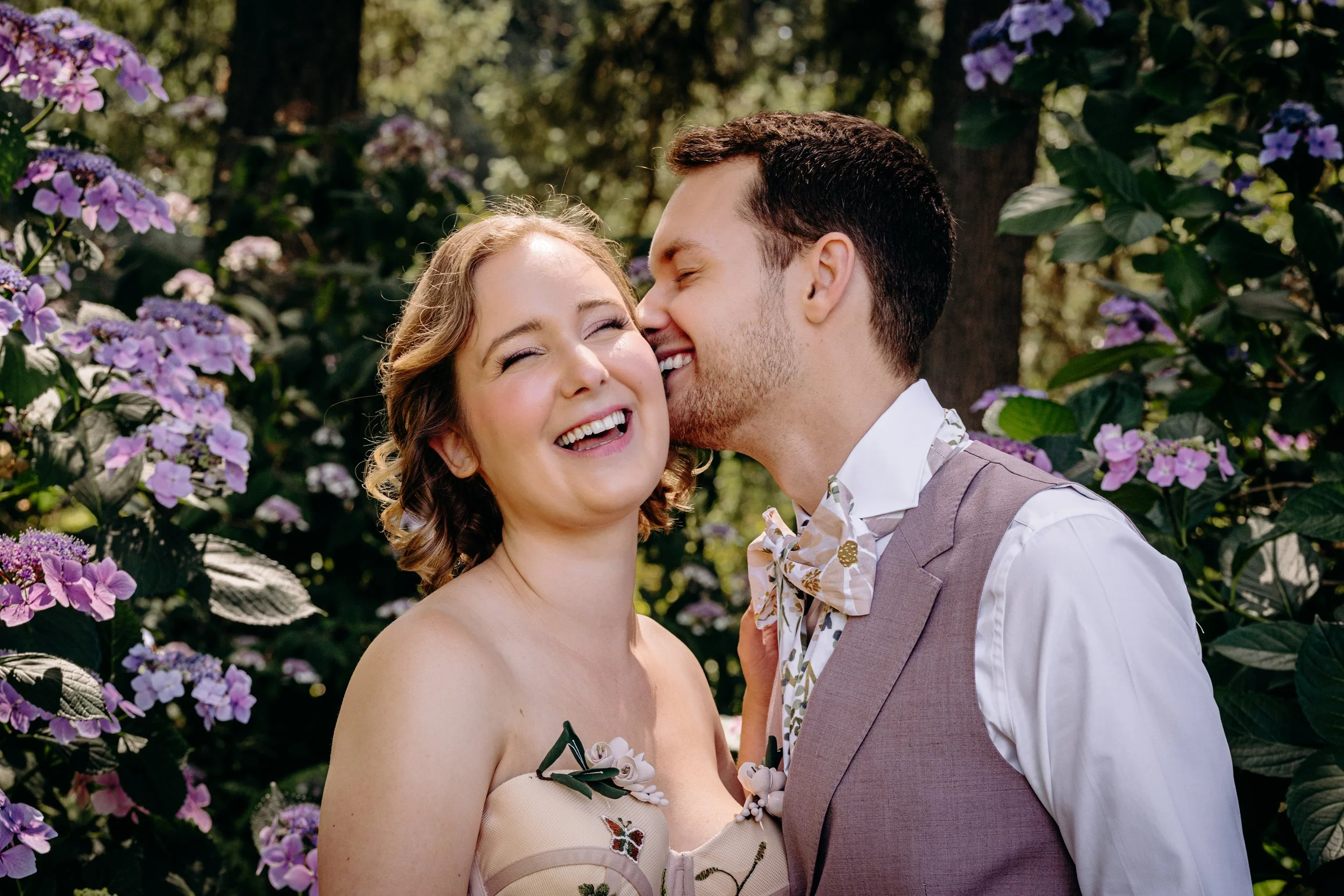A couple in love sharing a joyful moment with the man kissing the woman's cheek amidst purple hydrangeas and greenery.