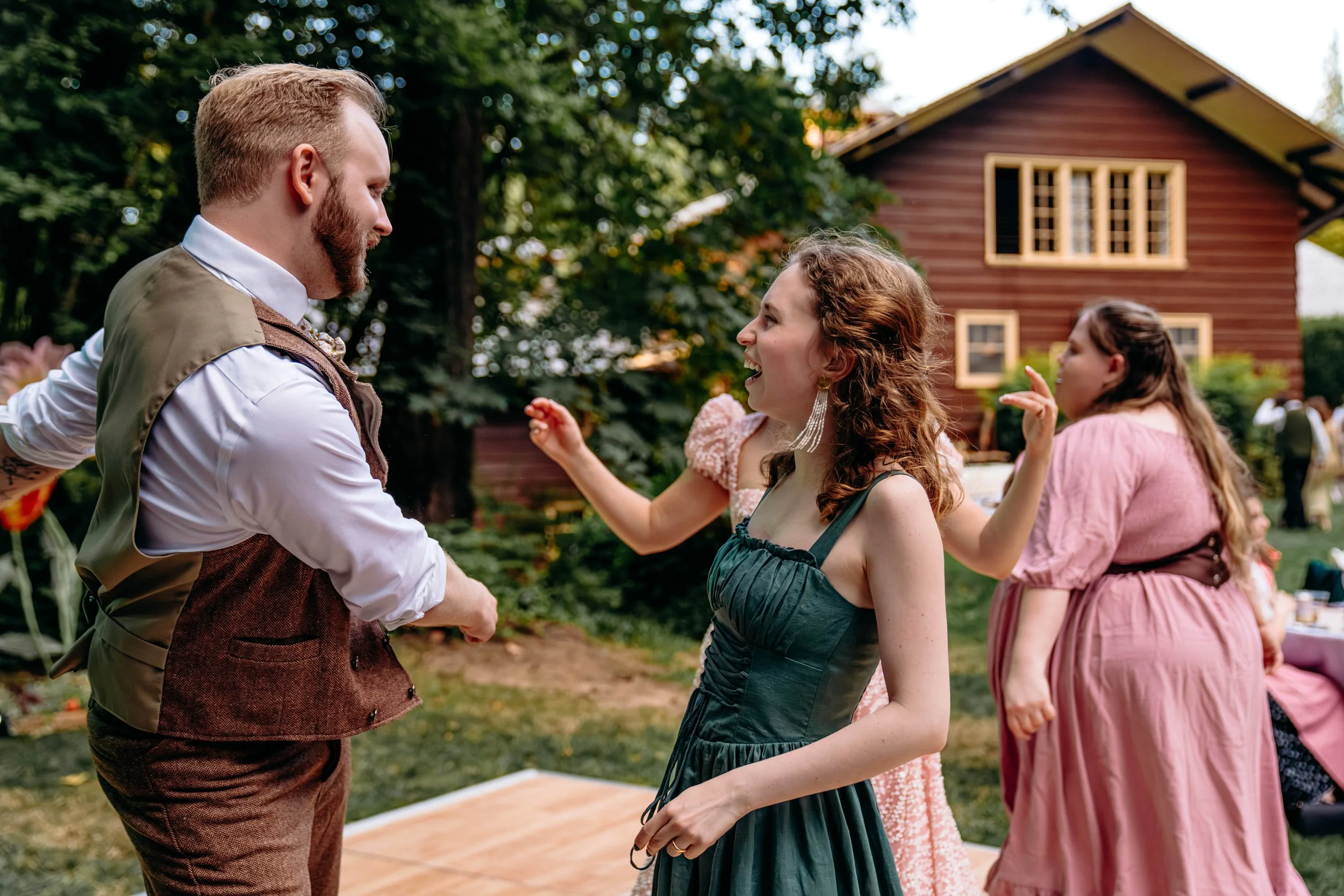 People dancing and socializing outdoors near a red wooden house in a garden setting.