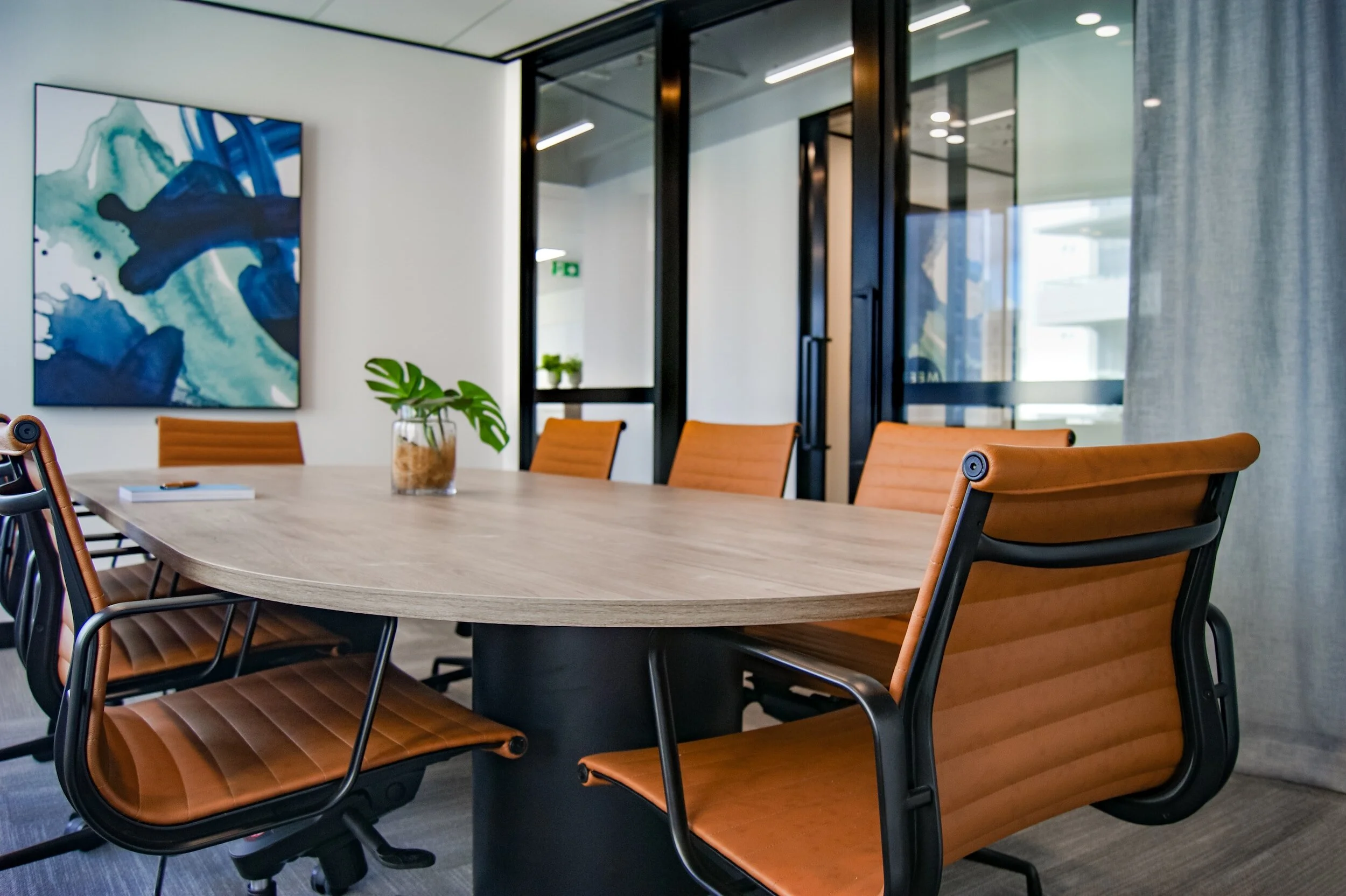 Modern conference room with a wooden table, six tan leather chairs, a abstract wall art, a houseplant in a glass container, and glass doors leading to another room.
