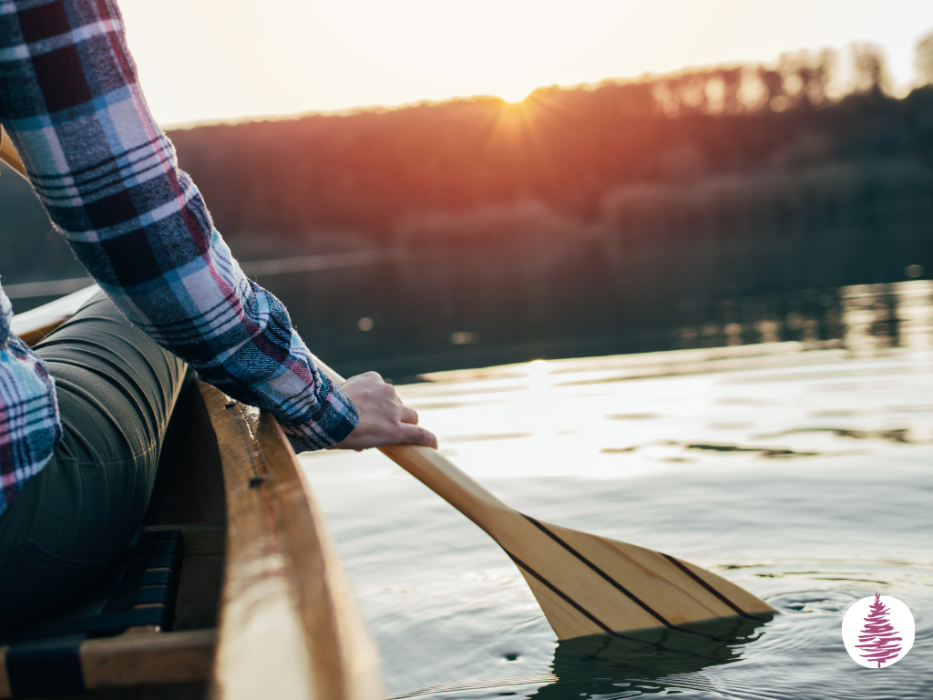 Seen from behind, a woman in a canoe dips her paddle into a smooth lake while the sun rises from behind the trees on shore.