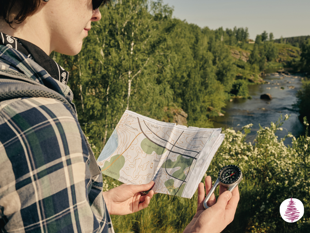 A person stands overlooking a river checking a compass against a map