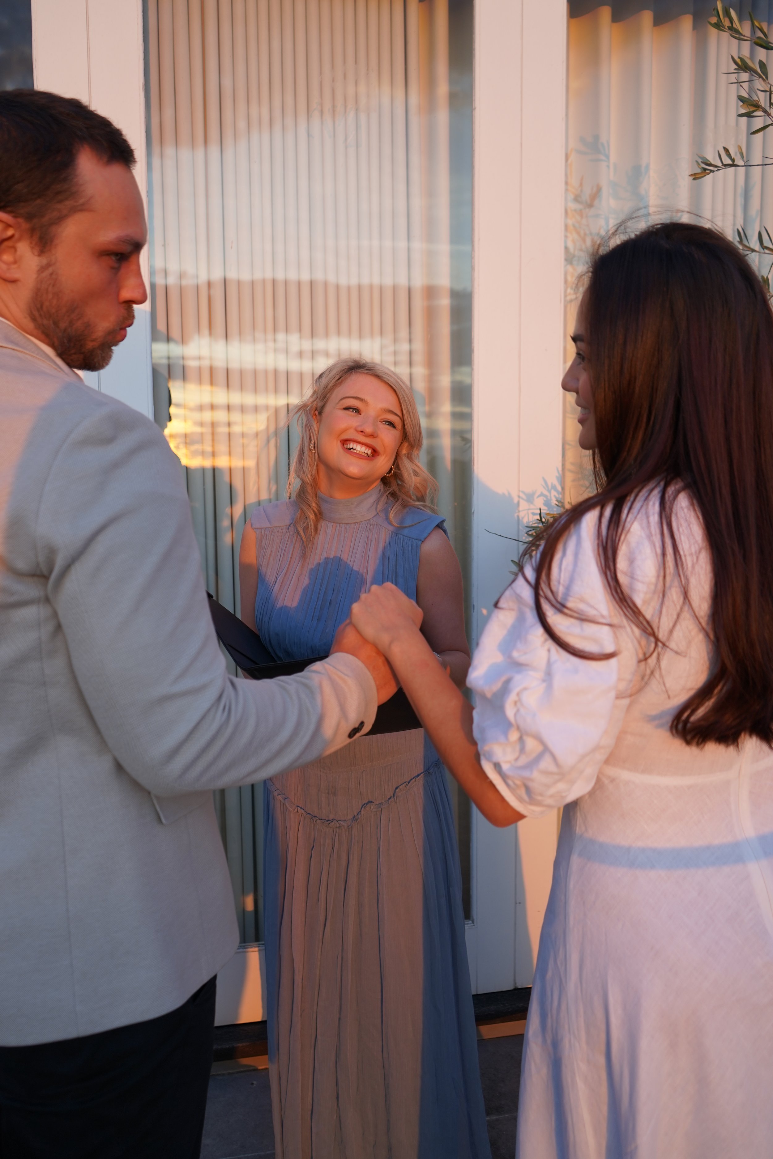 A group of three people shakes hands outside in front of a glass door during sunset, with a woman smiling in the background.