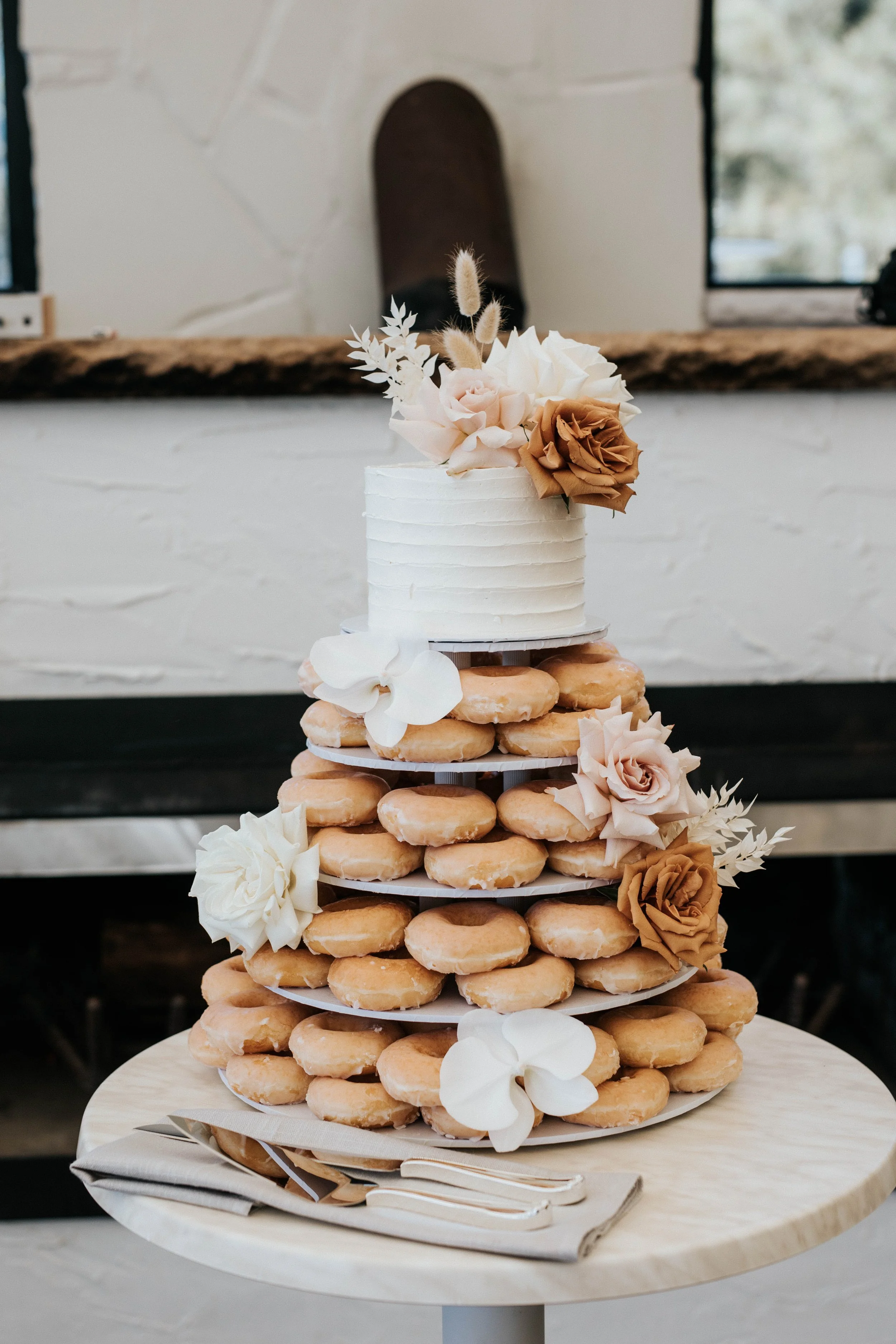 A multi-tiered dessert cake with a small white frosted cake on top, decorated with beige, white, and brown roses, white orchids, and dried flowers, placed on a layered stand of donuts, on a round wooden table with napkins and utensils, in a rustic setting.