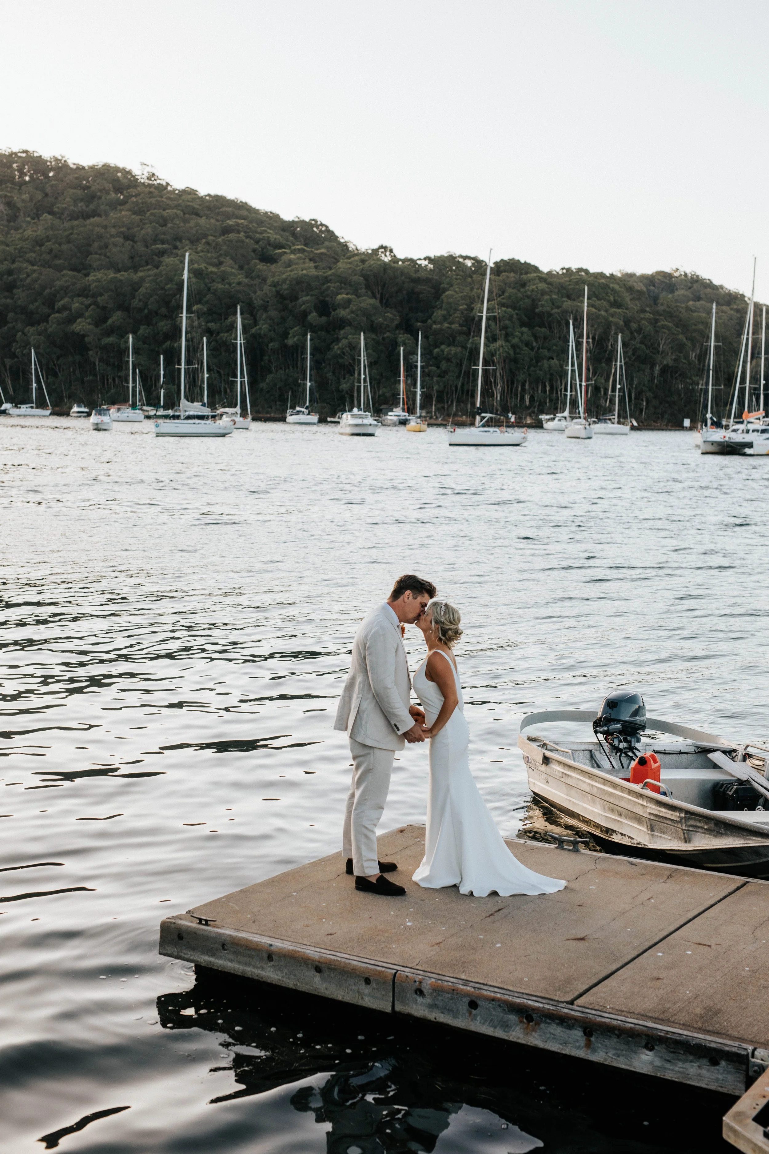 A couple dressed in wedding attire holding hands and touching foreheads on a dock by the water, with sailboats anchored in the background and a forested hillside across the water.