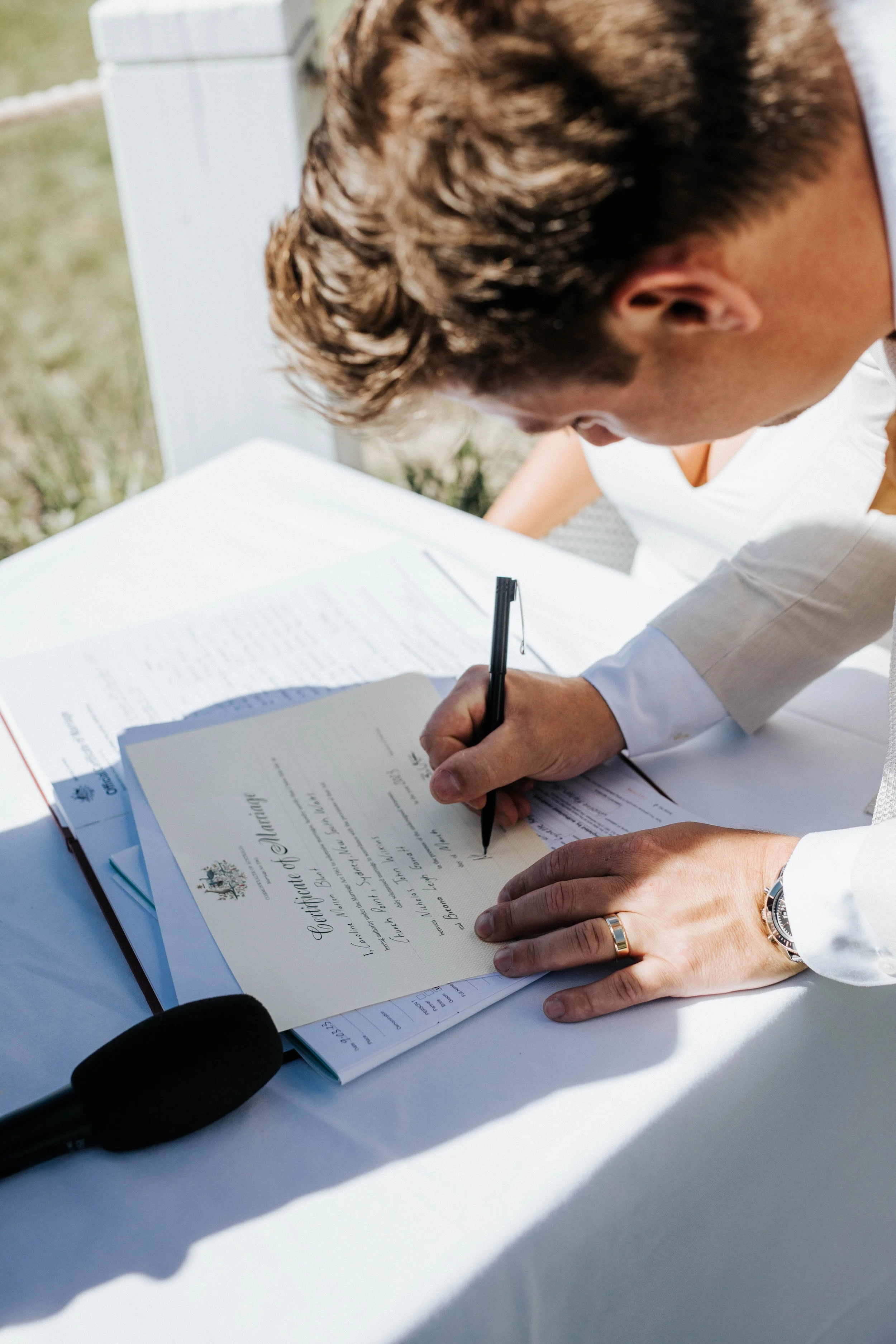 A man signing a document at a table outside, with a wedding certificate visible on top of the papers.