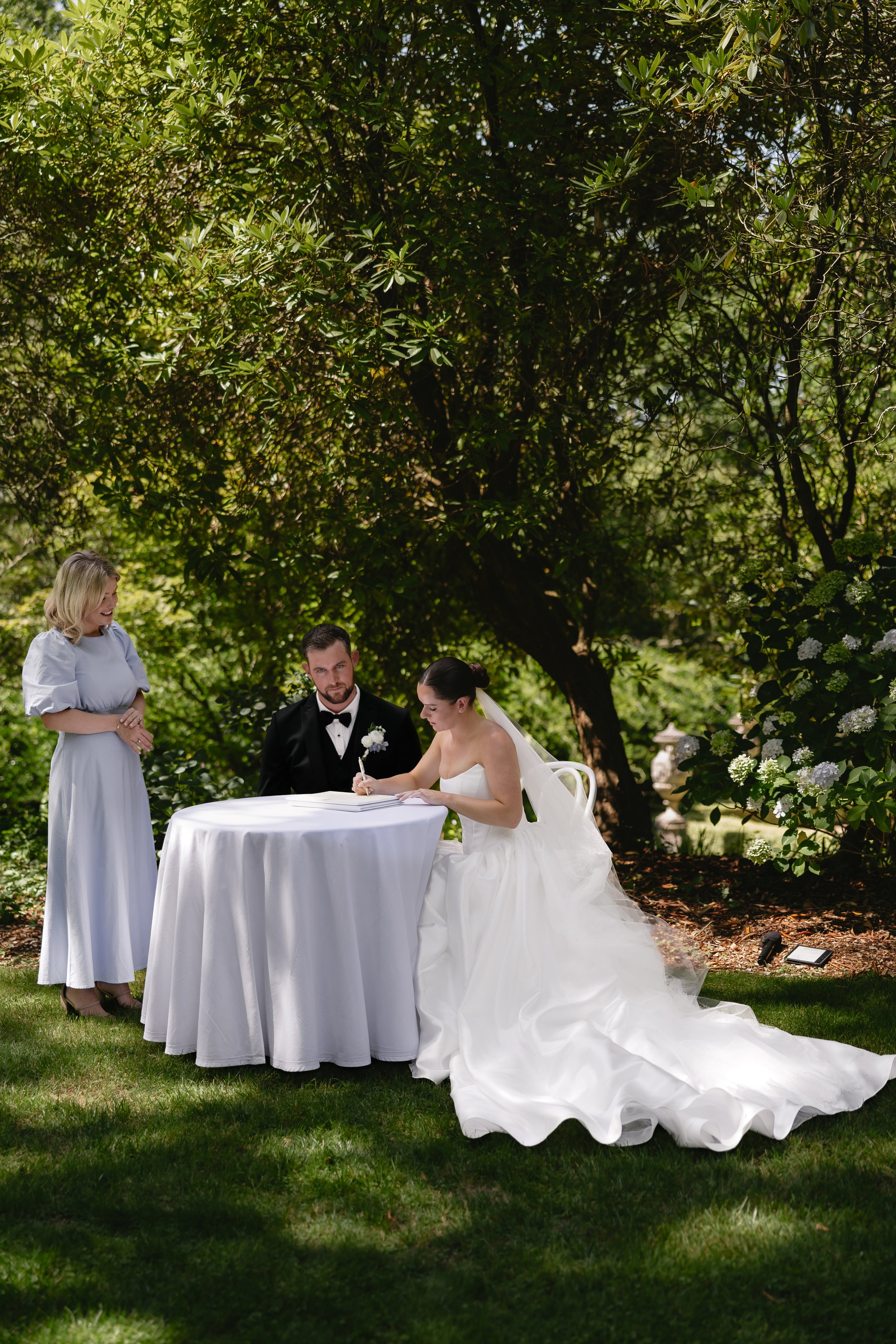 A couple dressed in wedding attire holding hands and touching foreheads on a dock by the water, with sailboats anchored in the background and a forested hillside across the water.