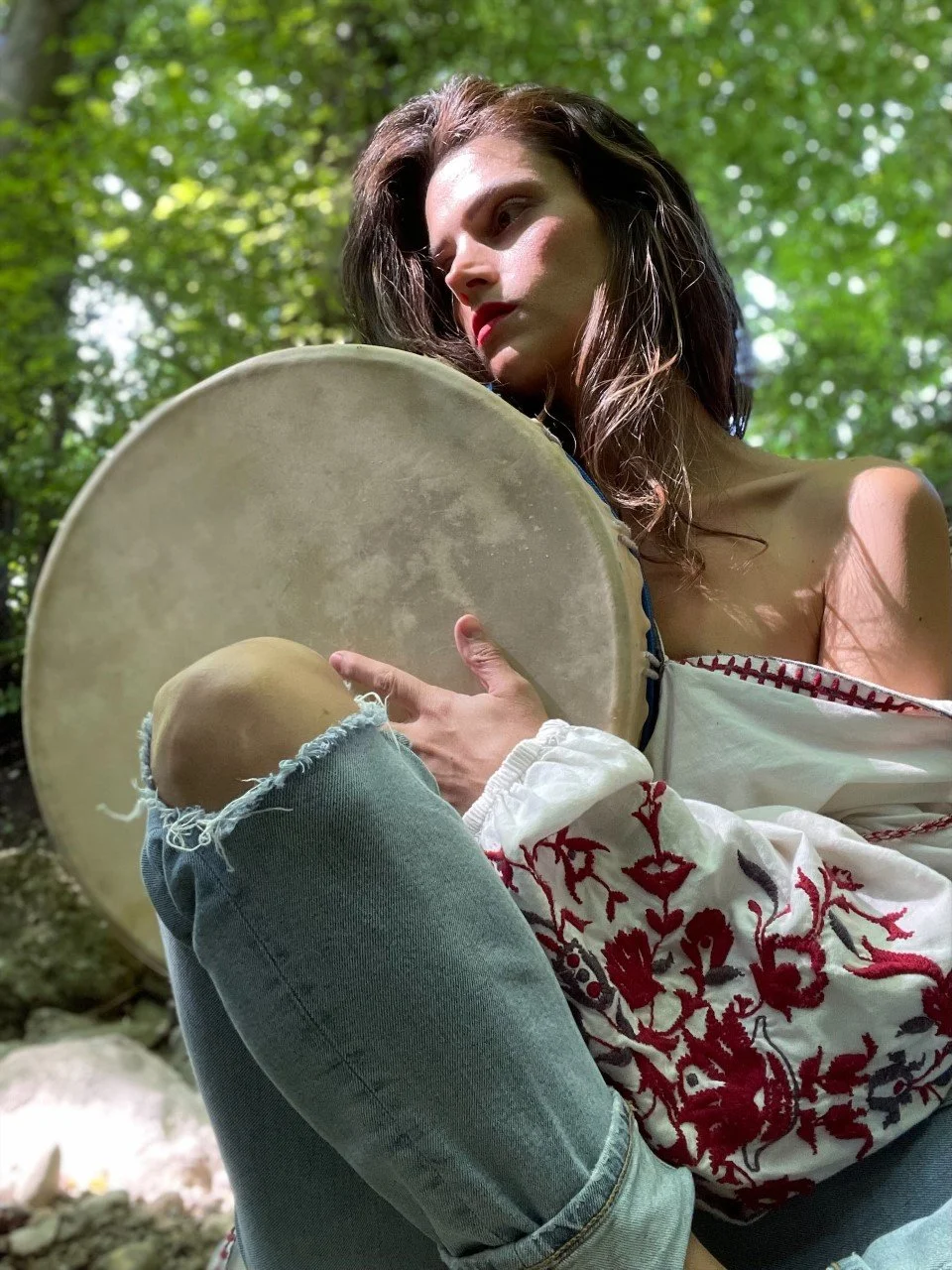 A woman with brown hair sitting outdoors in a forest, holding a large drum. She is dressed in a white off-shoulder top with red embroidery and ripped jeans. The sunlight filters through the green trees, creating a dappled light effect.