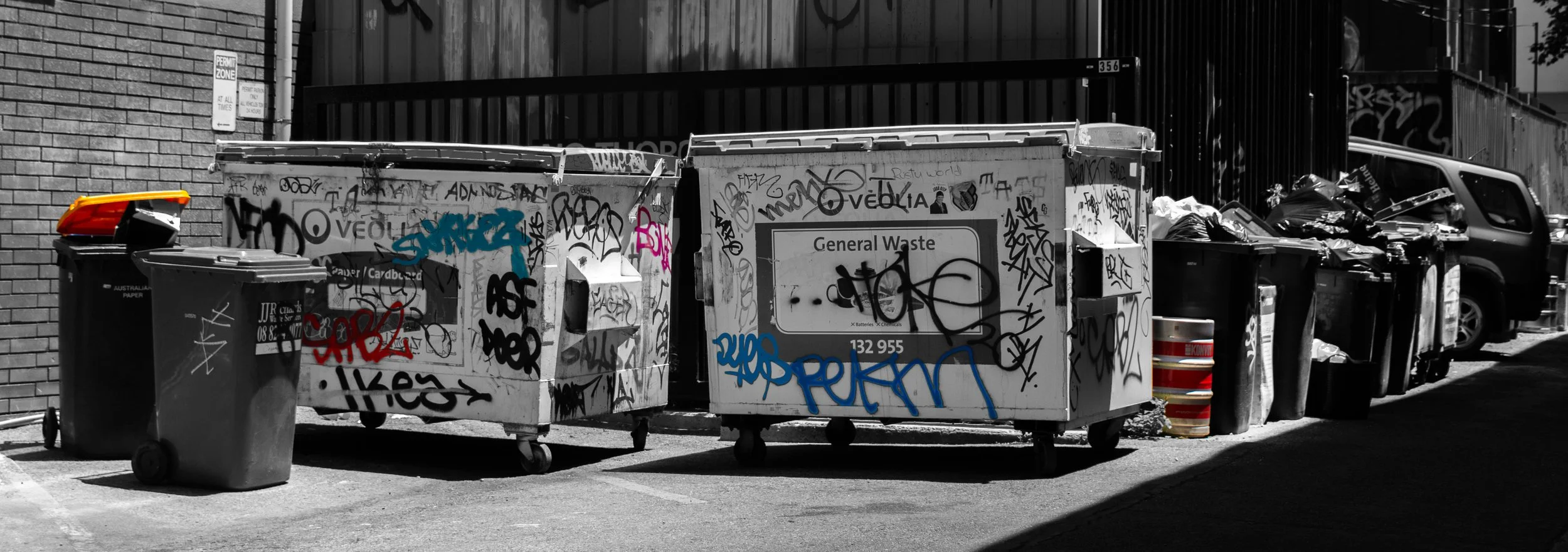 Multiple graffiti-covered trash dumpsters and bins in an alleyway, with a parked vehicle in the background.