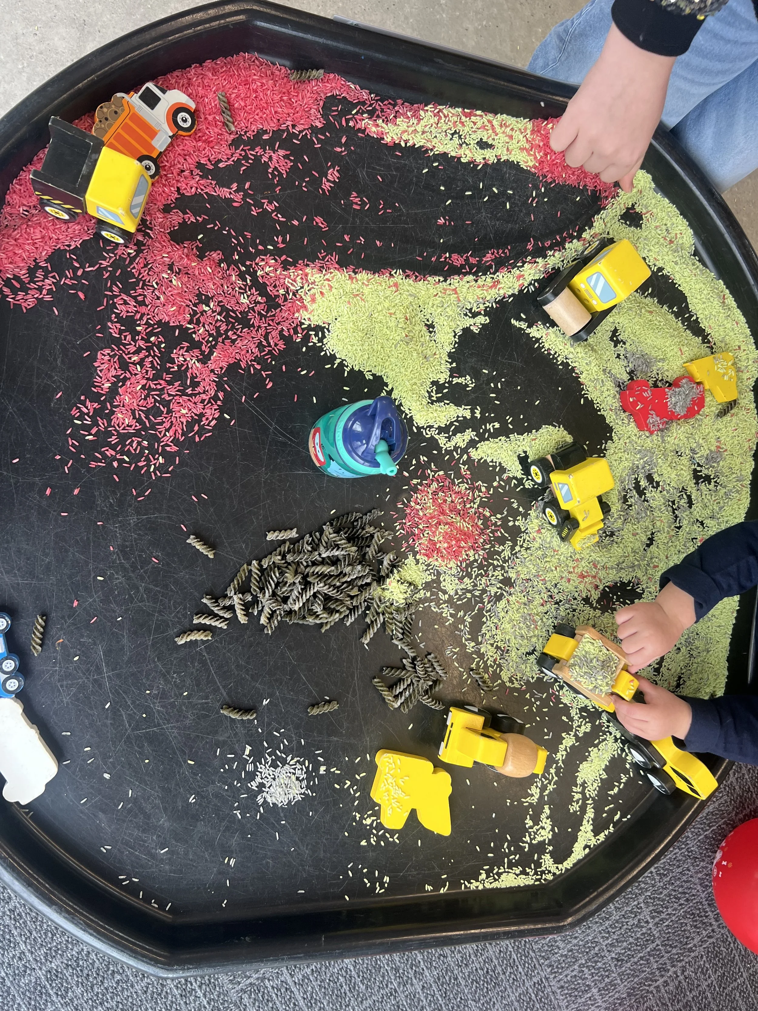 Children playing with toy trucks, rice, and pasta on a black tray shaped like a map of Asia.