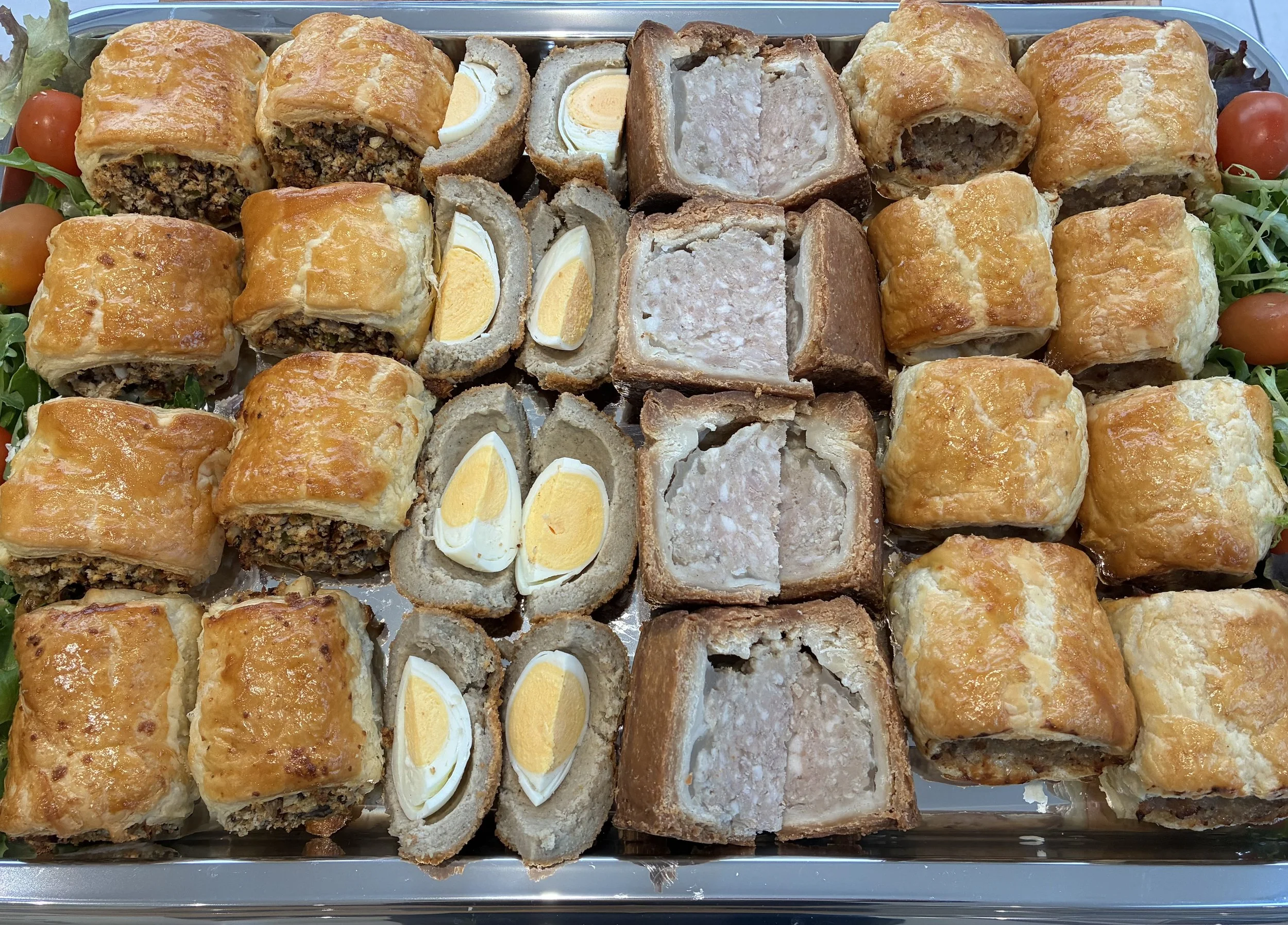 Assorted baked appetizers including sausage rolls, Scotch eggs, and pastry-wrapped meatloaf on a tray with cherry tomatoes and greens.