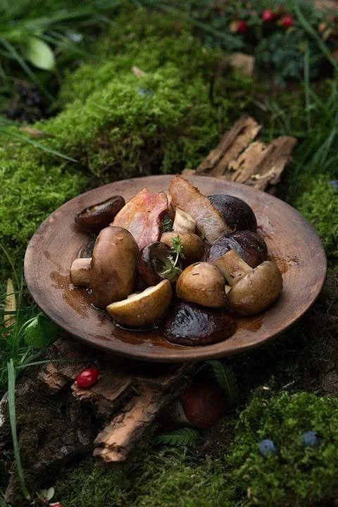 A wooden plate with assorted cooked mushrooms and bacon slices, placed on a mossy forest ground with green plants and small flowers.