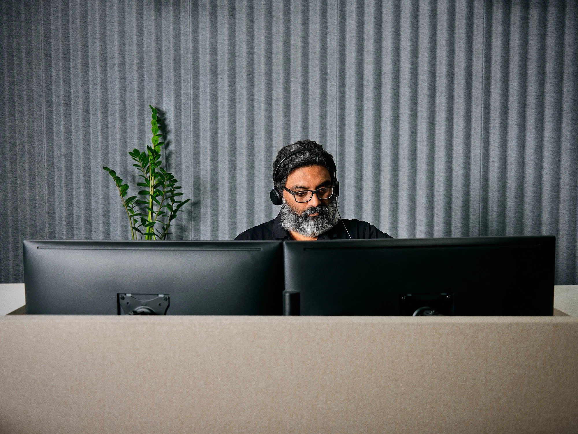 A man with glasses and a beard wearing a headset working at a desk with two monitors. There is a green plant on the left and a gray textured wall in the background.
