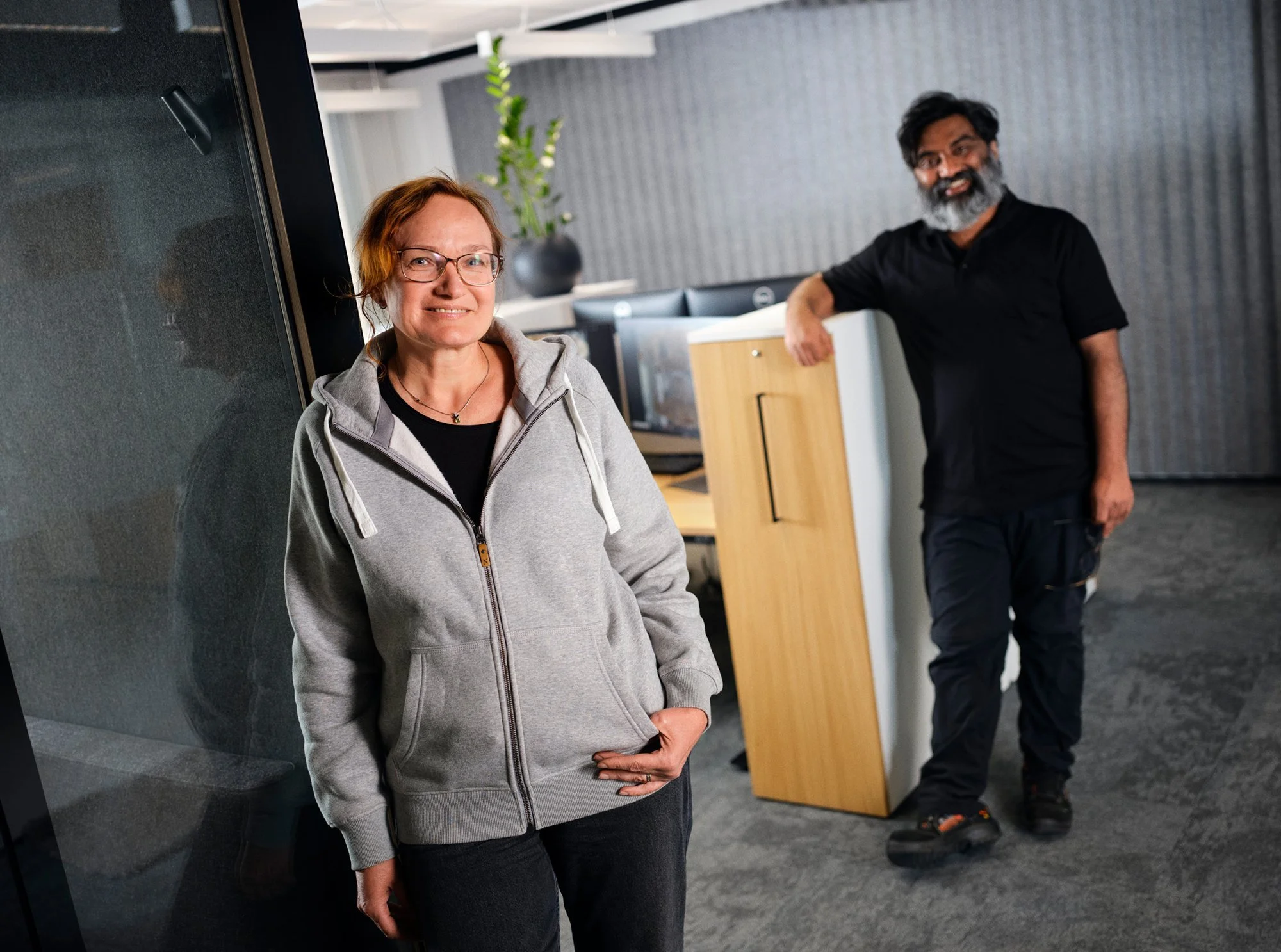 A woman with red hair and glasses wearing a gray hoodie standing next to a glass wall, smiling at the camera, with a man with dark hair and a beard in a black shirt and pants leaning against a wooden podium in a modern office space.