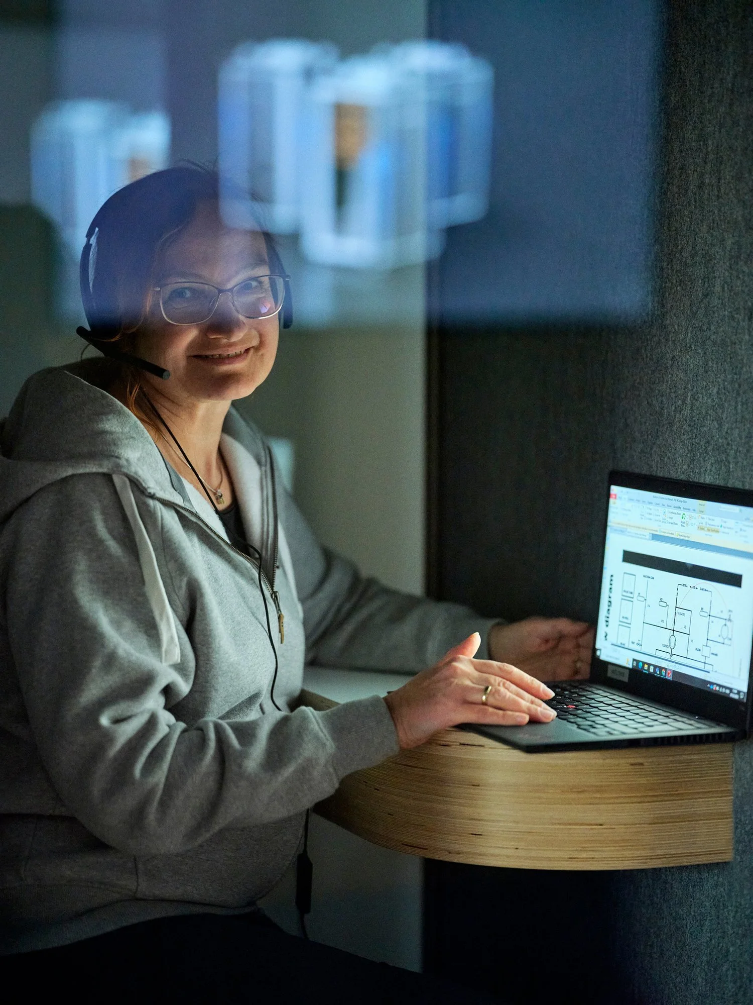 A woman wearing glasses and a headset sitting at a desk, looking at a laptop with a circuit diagram on the screen.