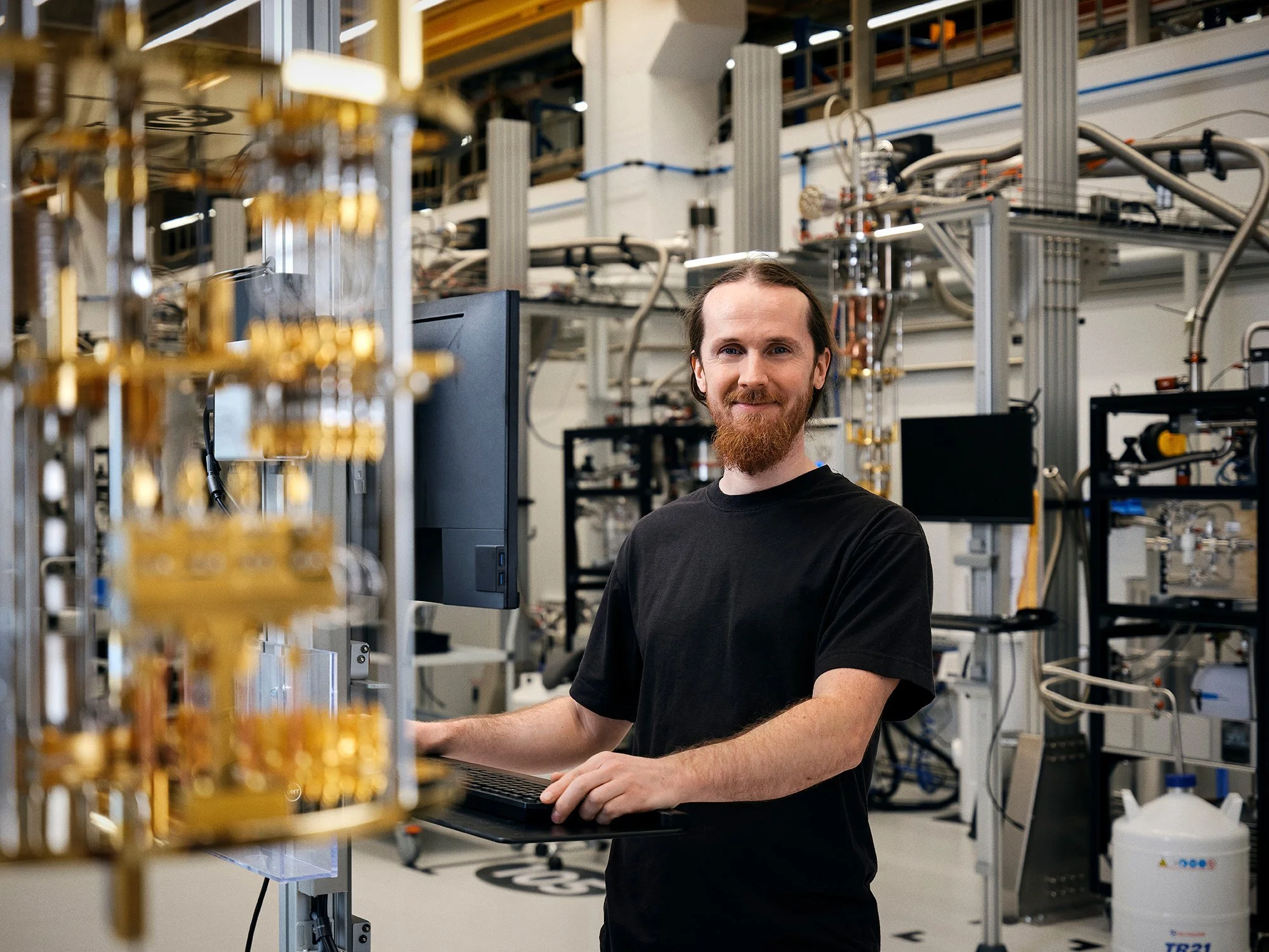 A man with a beard and long hair in a black t-shirt working on a computer in a scientific laboratory with complex equipment and machinery in the background.