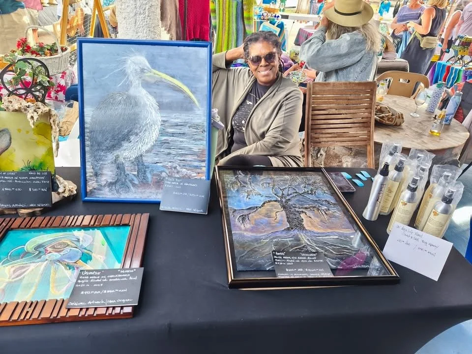 Artist sitting behind a table of handmade art at a Baja market, surrounded by framed pieces and natural light.
