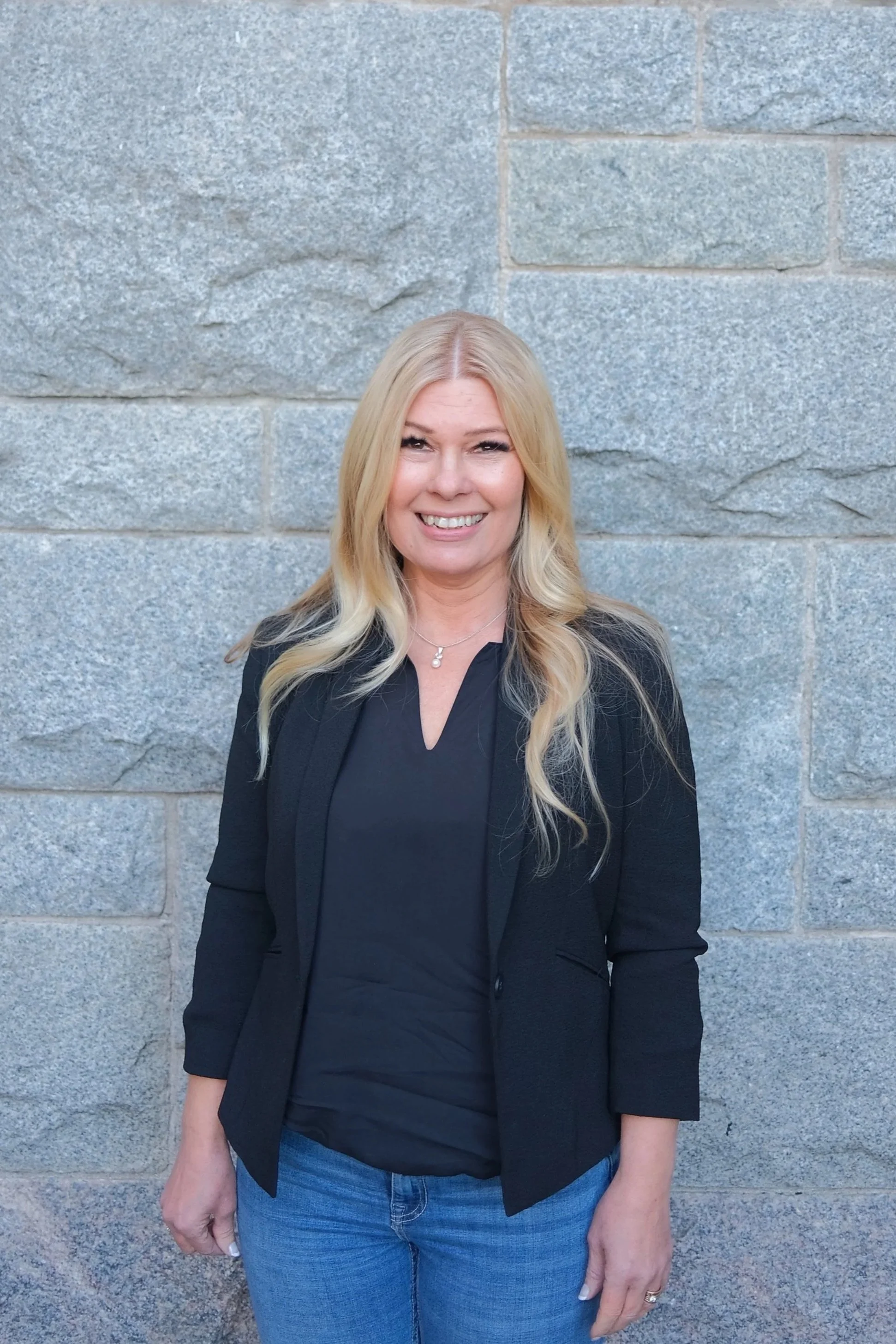 A woman with blonde hair smiling and standing against a stone wall, wearing a black blazer over a black top and blue jeans.