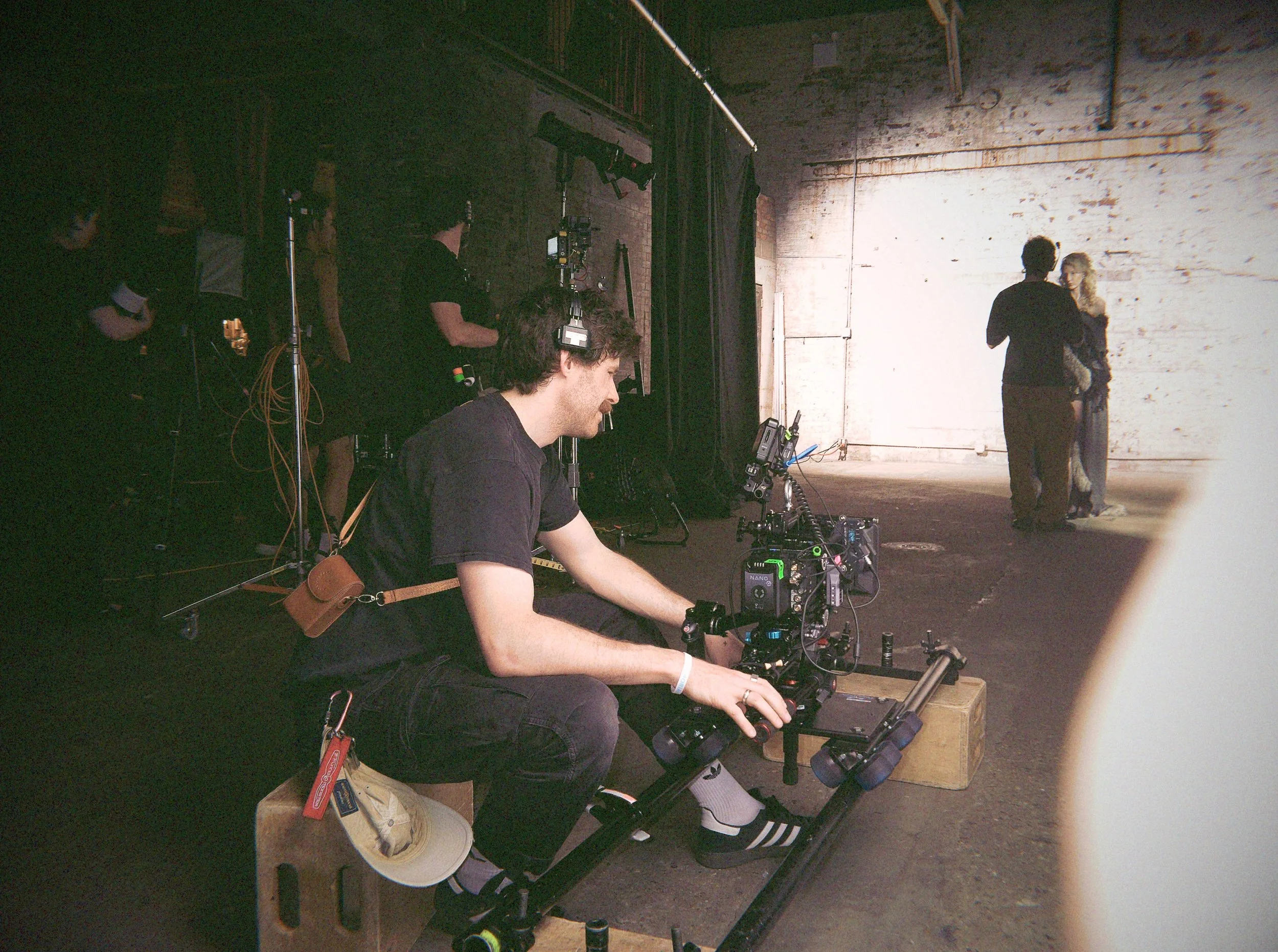 Filmmaking scene with a woman operating a camera on a tripod, crew members with equipment in a dimly lit industrial space, two actors in costume in the background, and various filming gear and accessories on a wooden table.