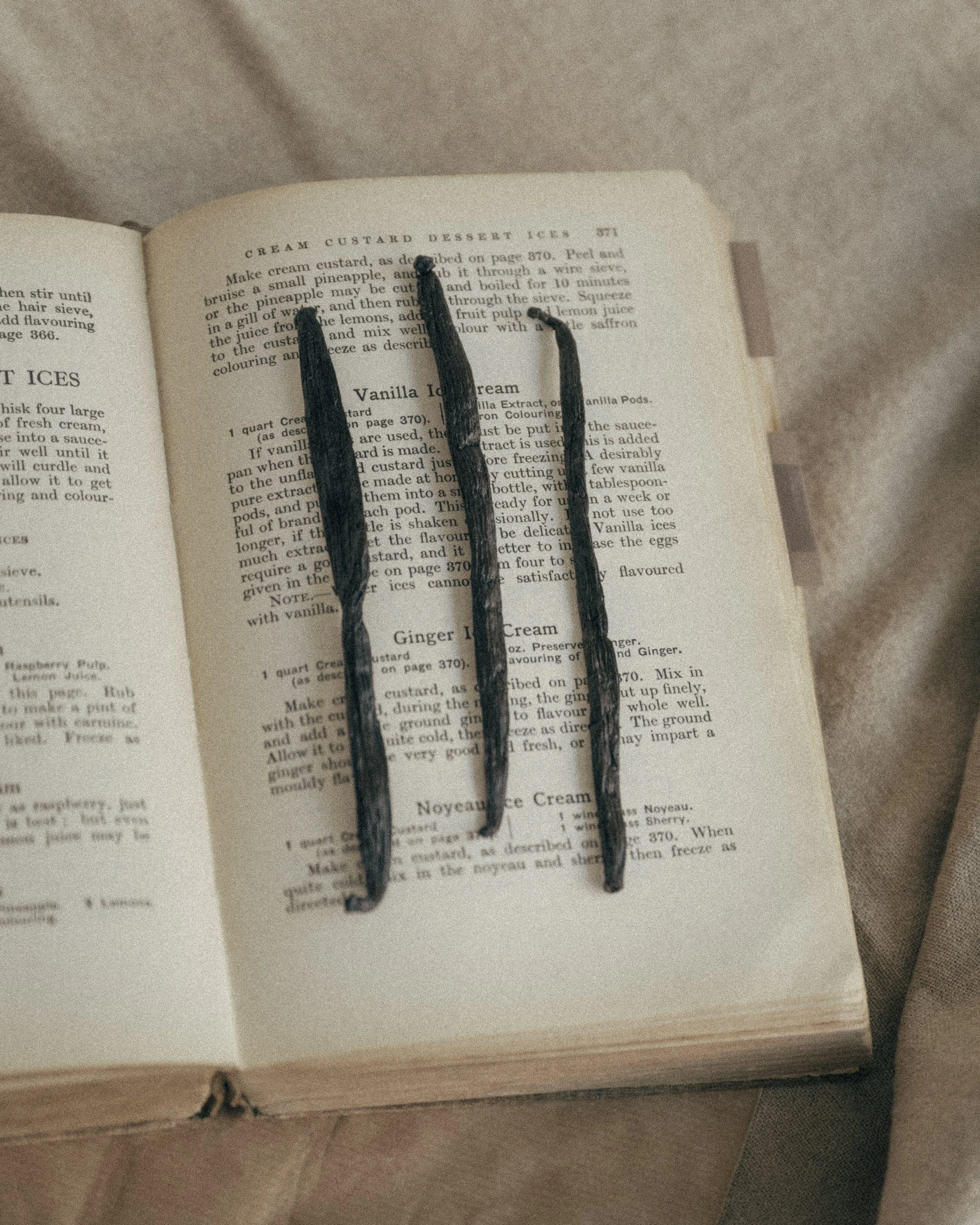 Three dark vanilla pods lying on an open vintage cookery book, on a page of old recipes for vanilla and ginger ice cream.