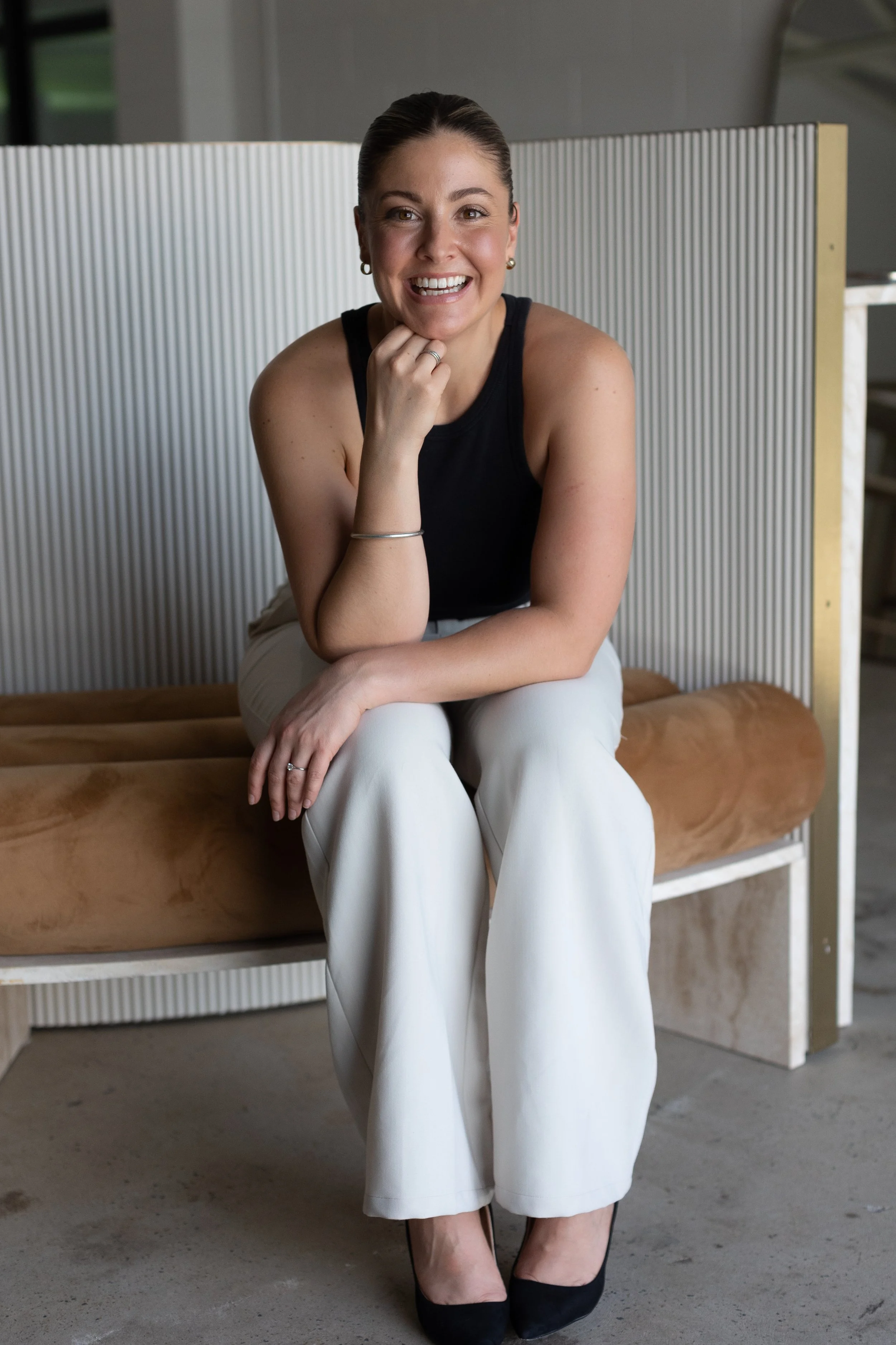 Jarrah Wallace - Kajabi Systems & Automations Partner. A woman with dark hair pulled back, wearing a black sleeveless top, white pants, and black shoes, sitting on a tan bench with a modern, textured white and gold backdrop in a neutral-colored room.
