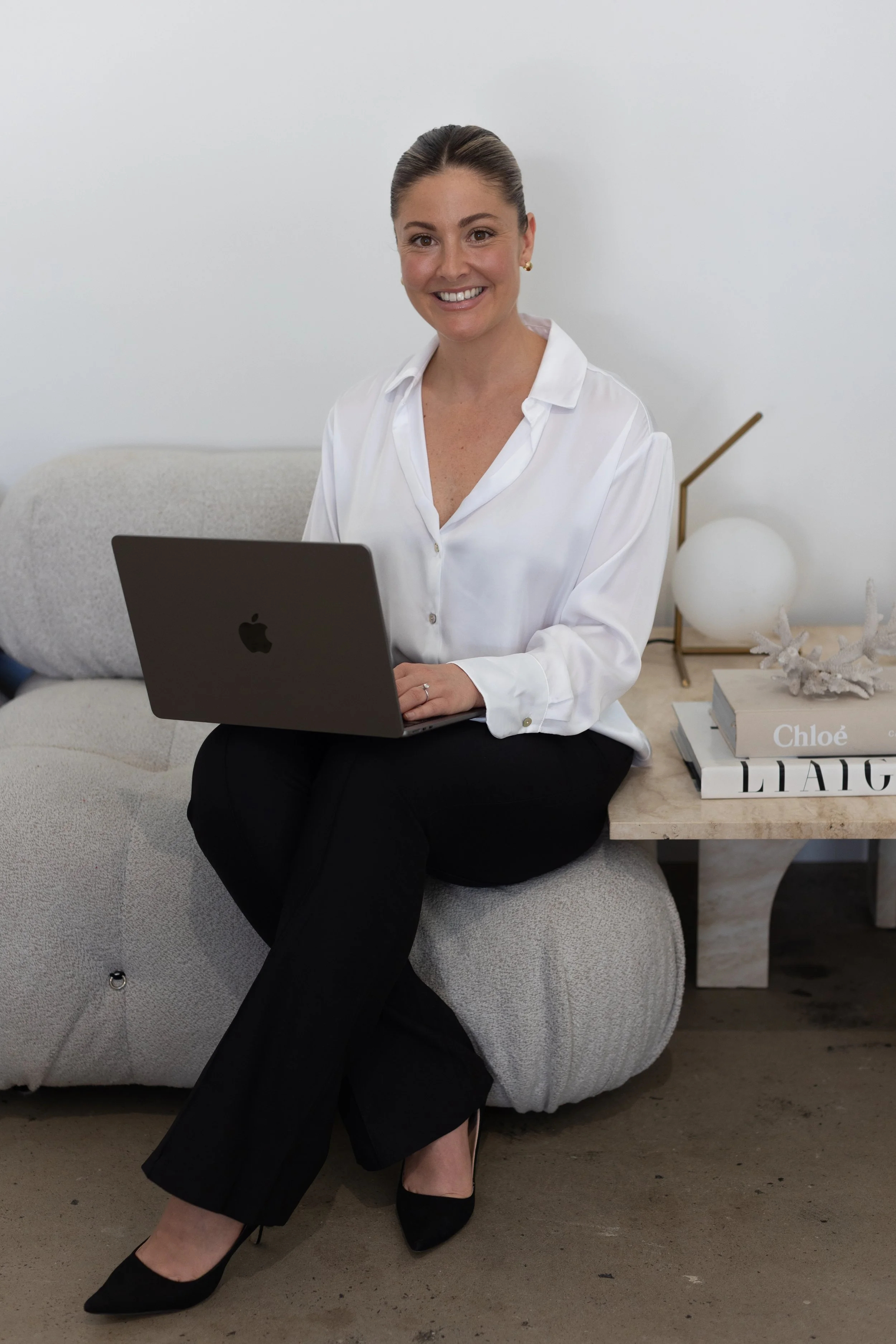 A woman sitting on a light-colored, plush ottoman in a modern, minimalistic room, working on a silver MacBook. She is smiling, wearing a white blouse, black pants, and black high heels, with her hair tied back. A decorative side table with books including titles 'Chloé' and 'LIAQ' and a modern lamp with a gold stand and white globe are in the background.