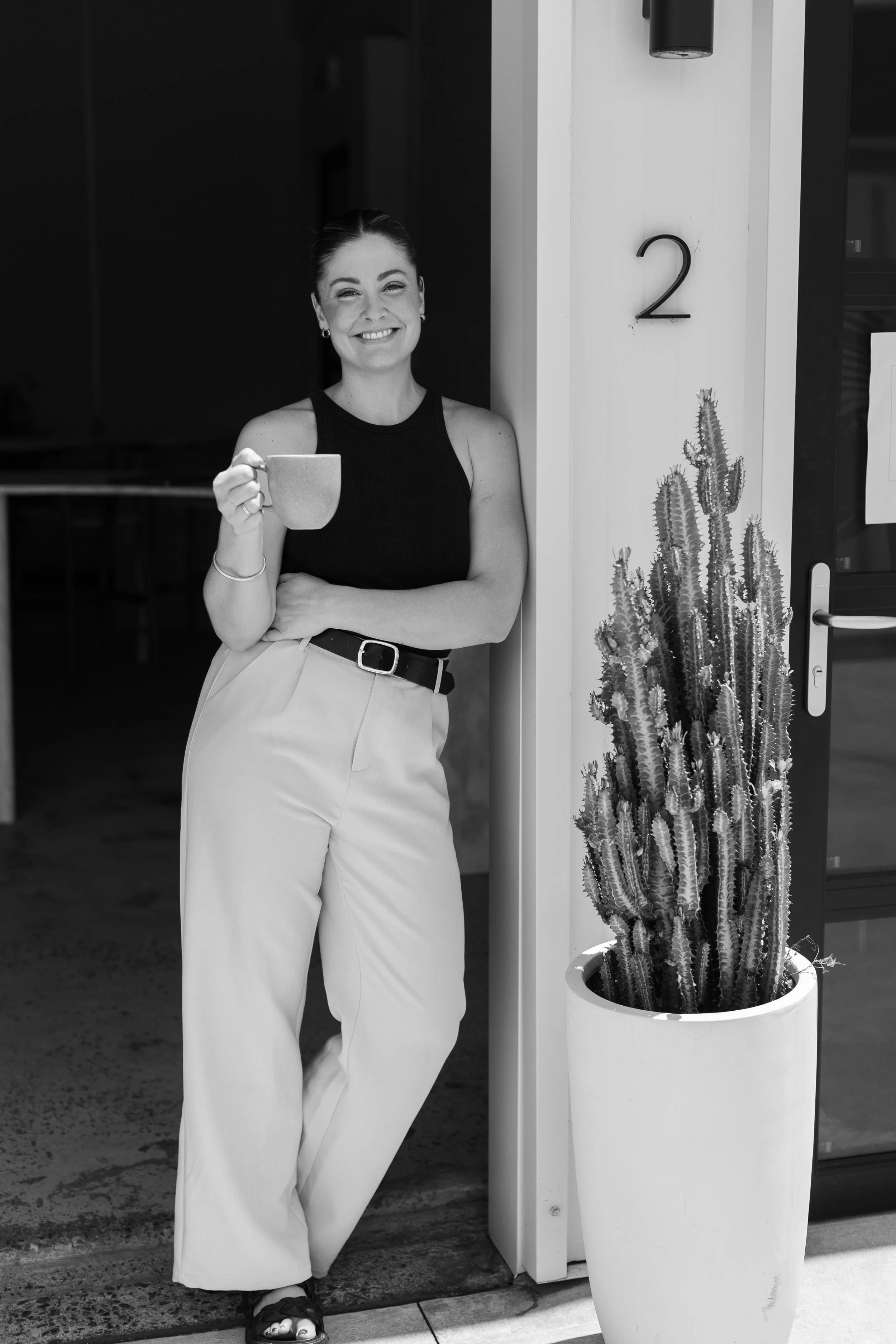 Jarrah Wallace - Kajabi Systems & Automations ParA woman standing near a cactus plant in a pot, holding a mug and smiling at the camera. She is wearing a sleeveless top and light-colored trousers, standing outside a building marked with the number 2.