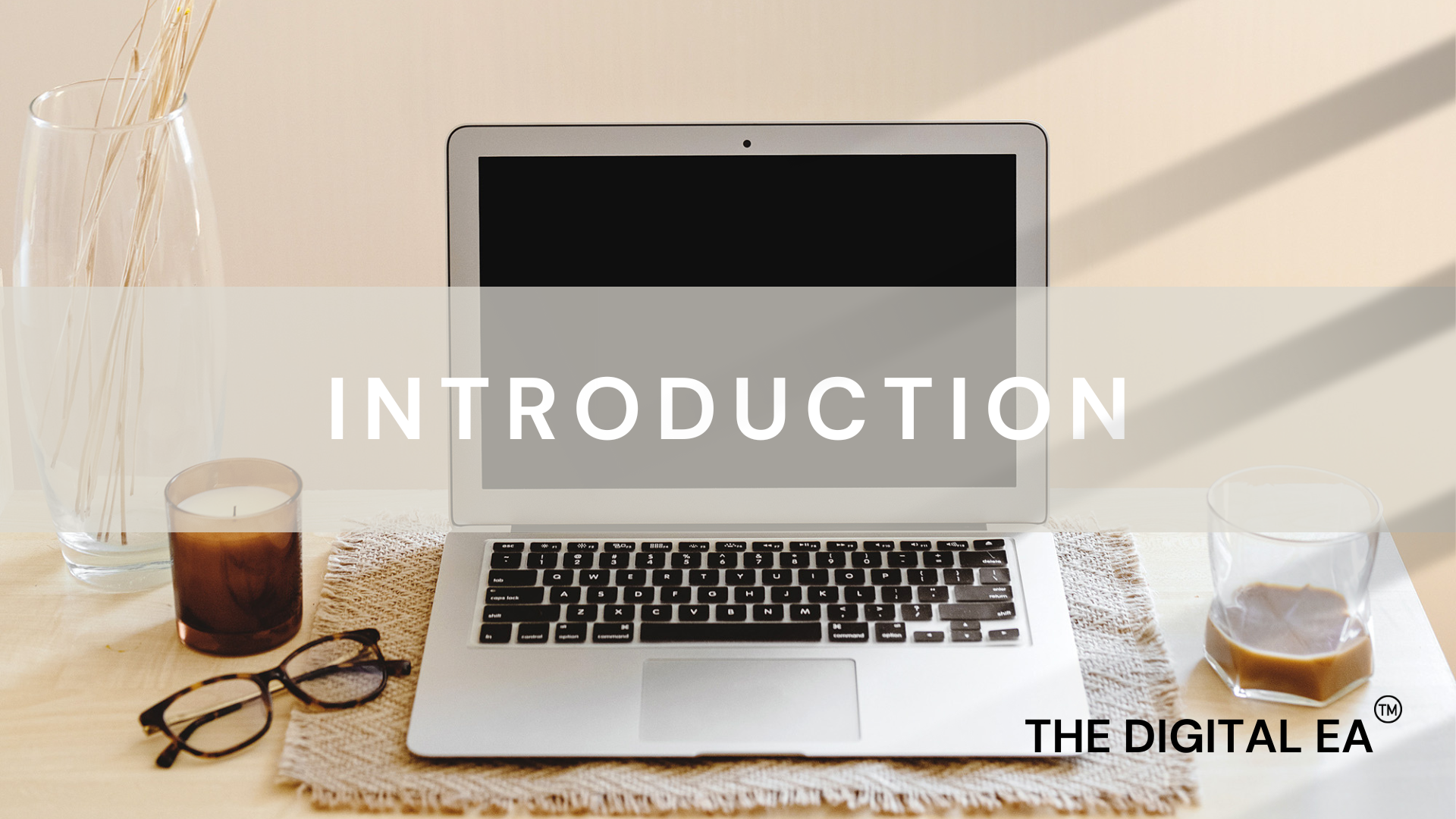 Jarrah Wallace - Kajabi Systems & Automations ParA MacBook laptop on a wooden desk with a glass of coffee, sunglasses, and a jar of decorative sticks, with the word 'INTRODUCTION' overlayed on the screen and 'THE DIGITAL EA' logo at the bottom right.