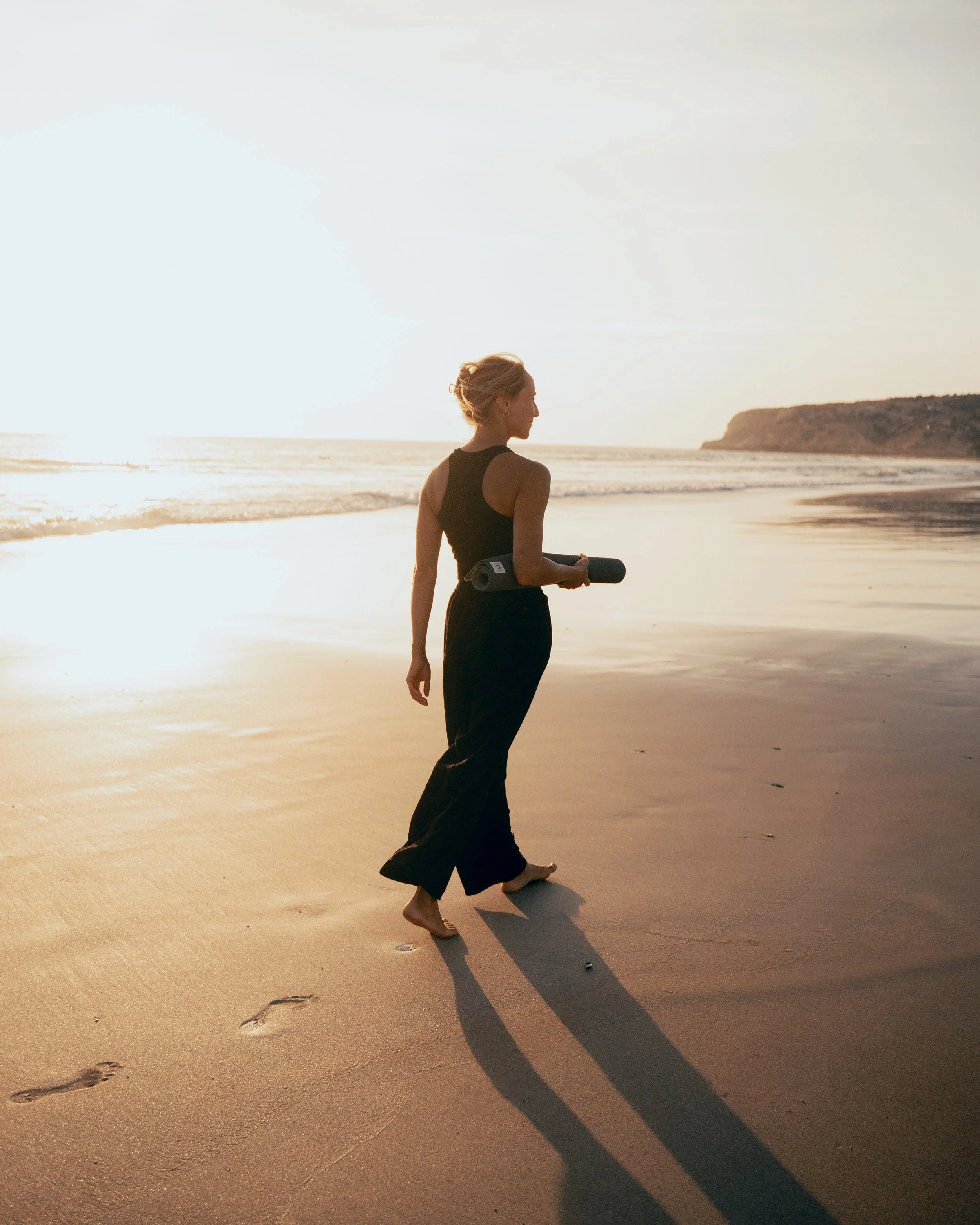 Woman walks down the Beach with a Yoga mat in her arms in golden sunset light