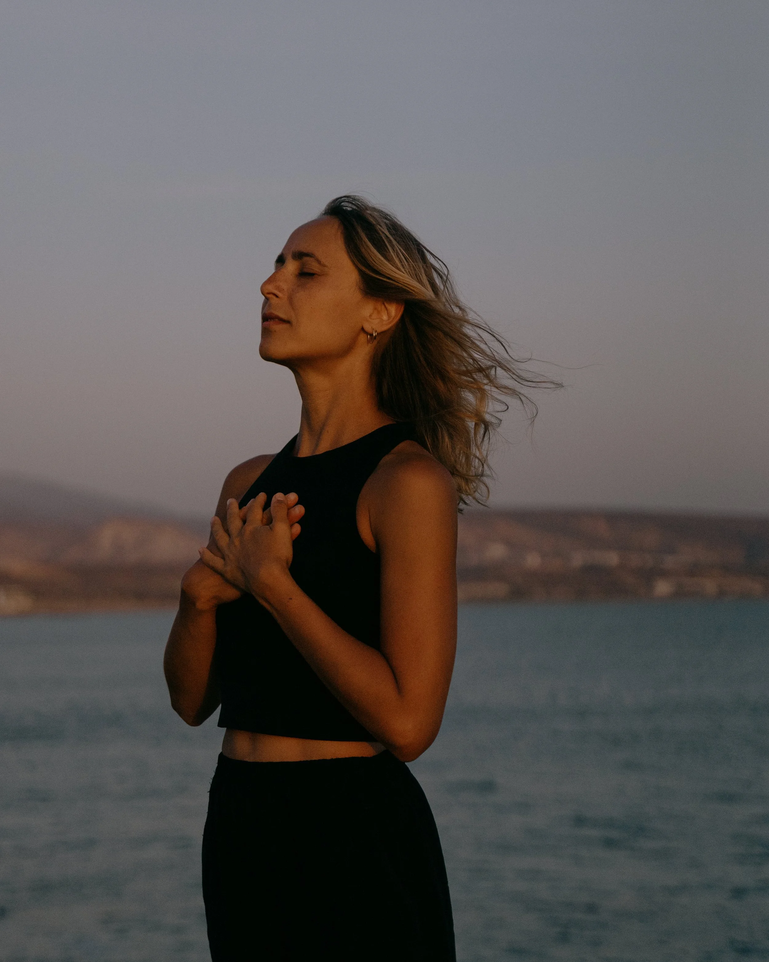 Woman practicing yoga or meditating outdoors near water, with her hands over her heart and eyes closed, during sunset.