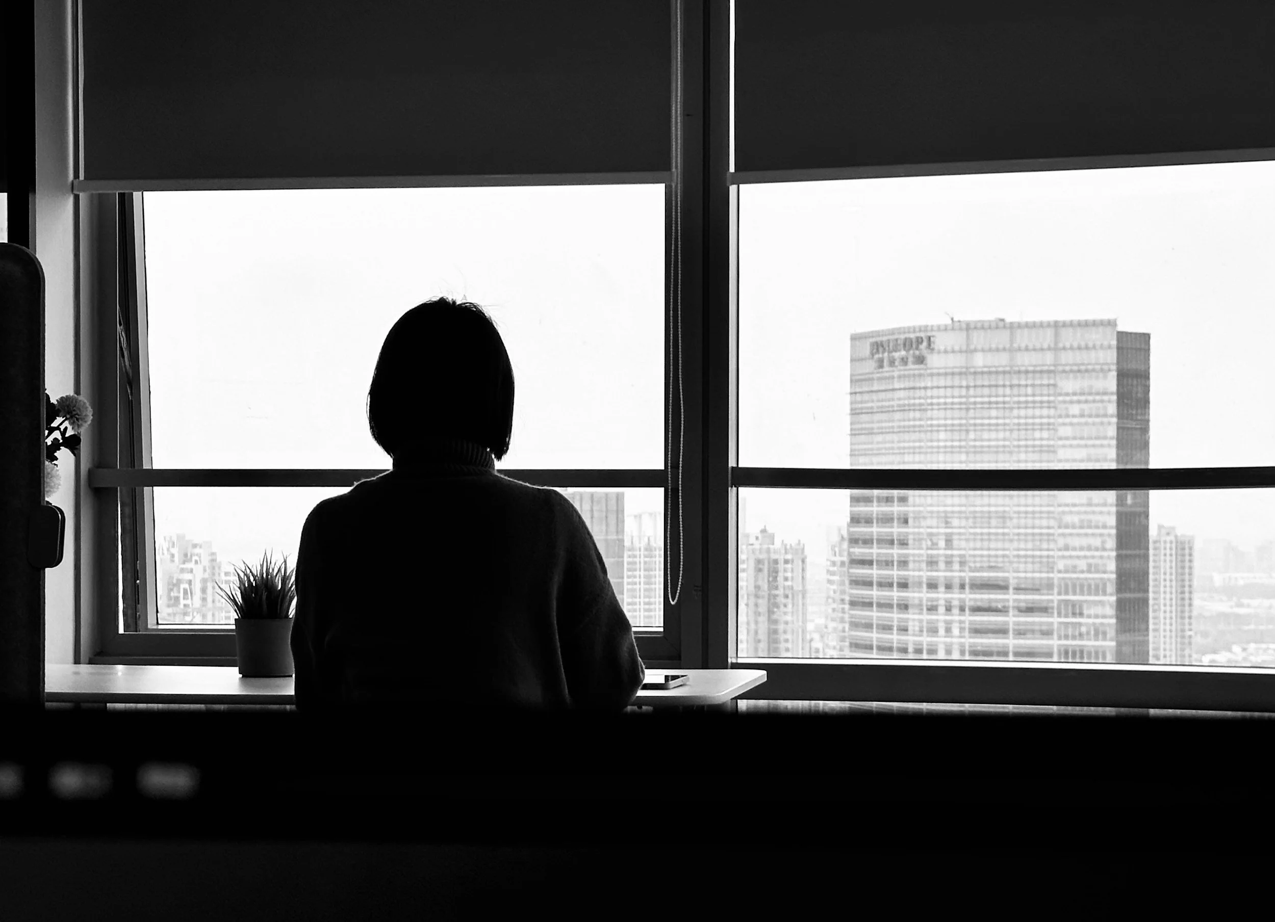 Black and white image of a person standing at an office window looking out onto a city with large buildings outside.