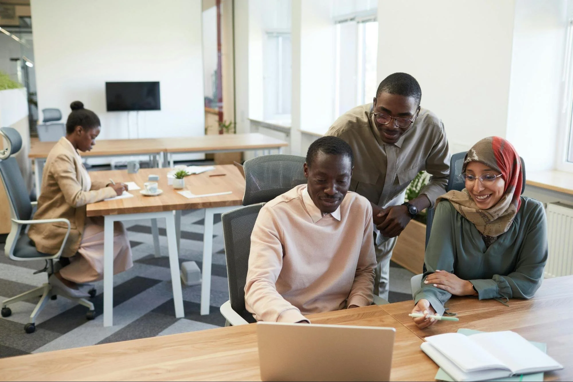 On the left in the background, a black woman sits by herself working at a desk. In the foreground on the right, two black men and a woman in a hijab face a laptop screen in an office setting.