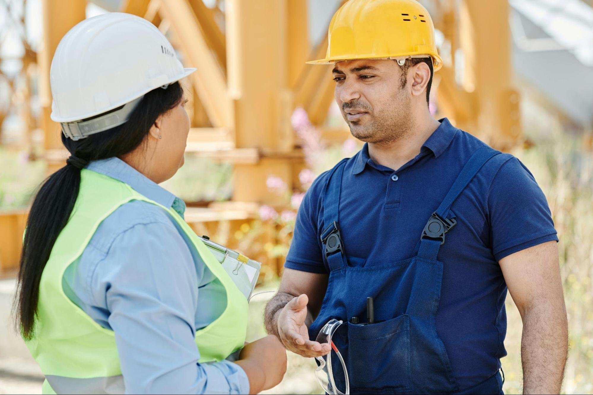 A woman’s back is on the left in a green safety vest and a construction hard hat. She faces a man on the right dressed in blue and wearing a yellow hard hat.