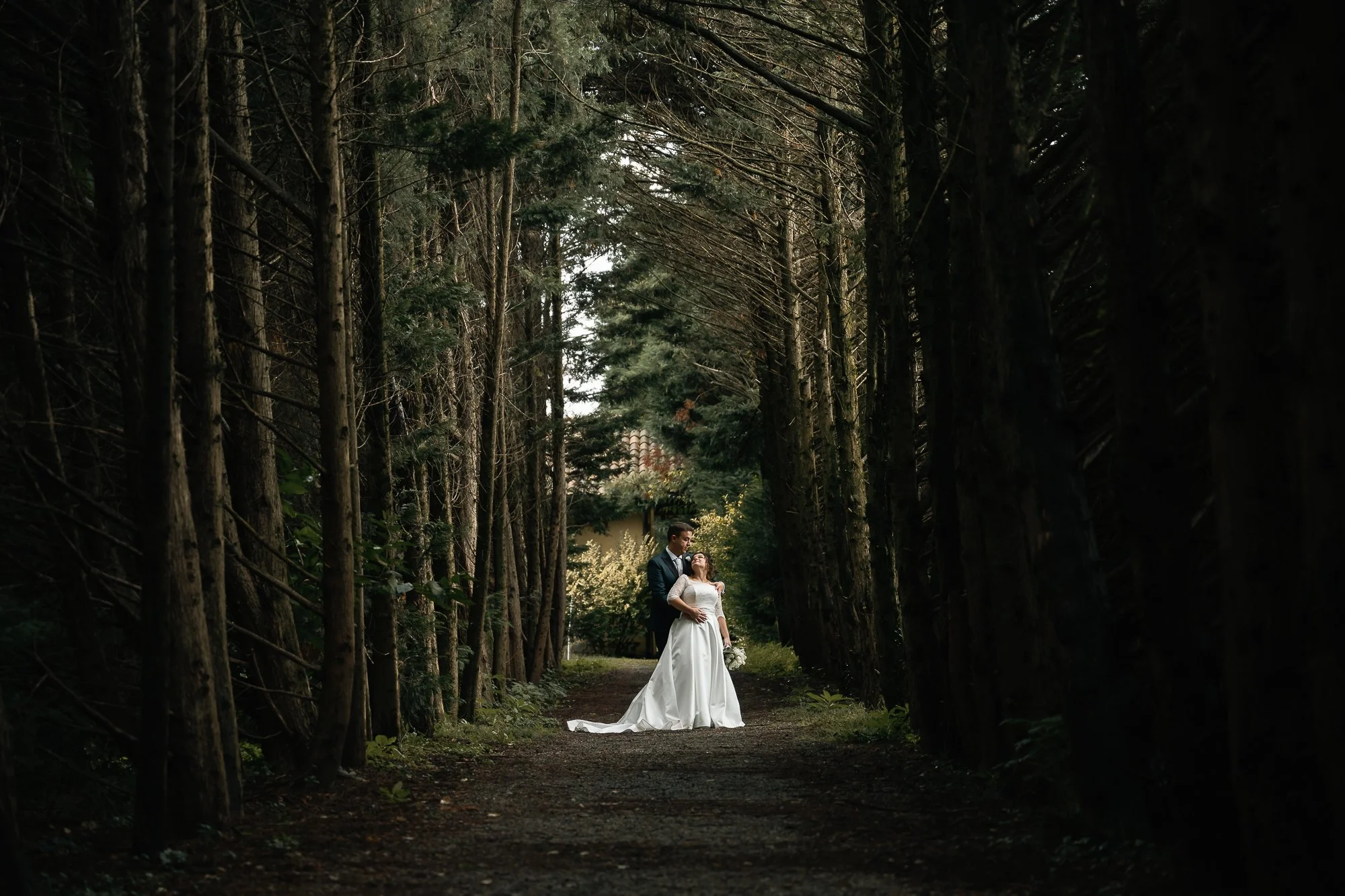 A bride in a white wedding gown and a groom in a dark suit standing on a path in a dense forest, embracing each other.