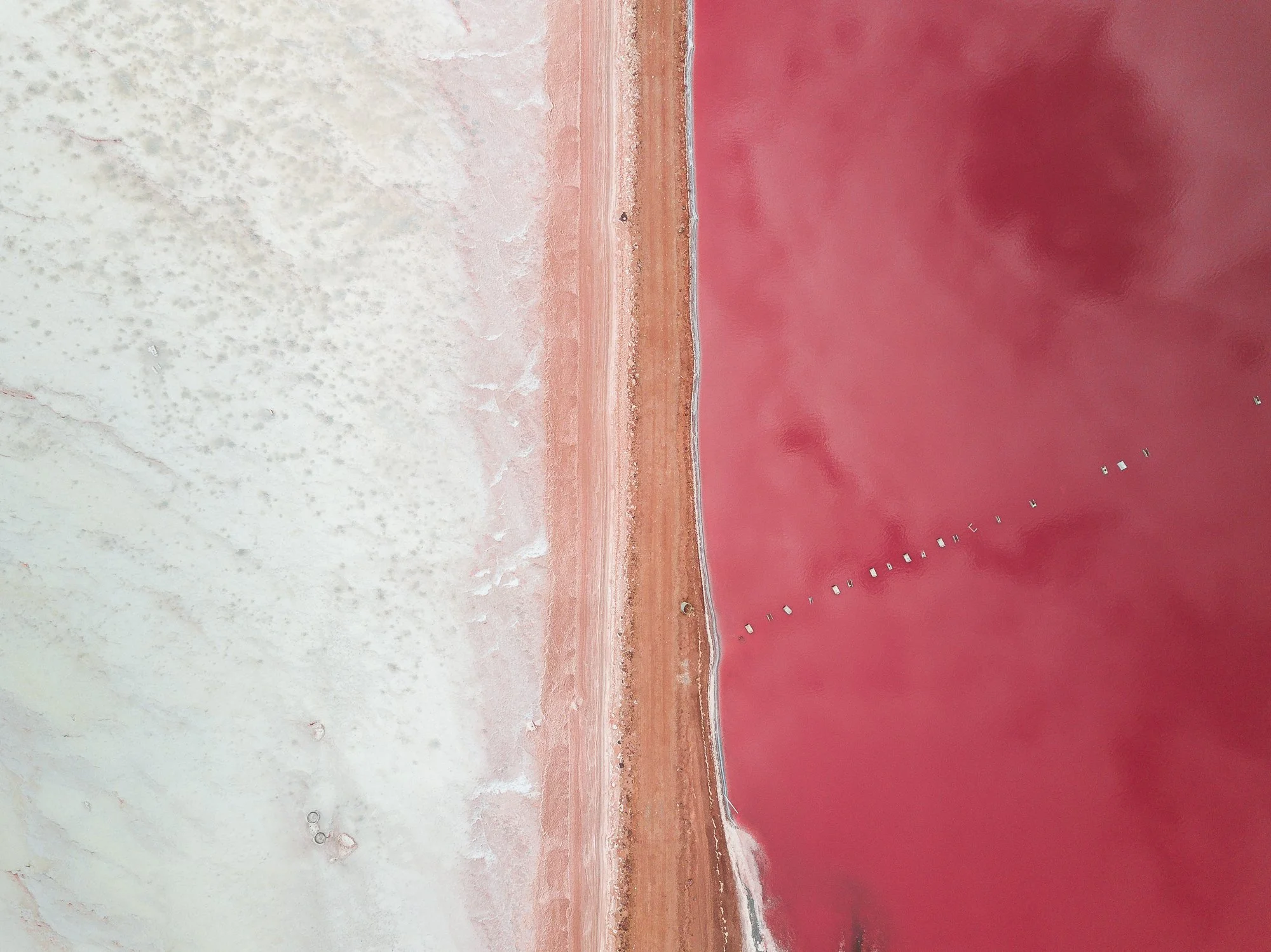 An aerial view of a pink salt pond divided into two sections by a narrow strip of land, with small structures or boats visible on the water.