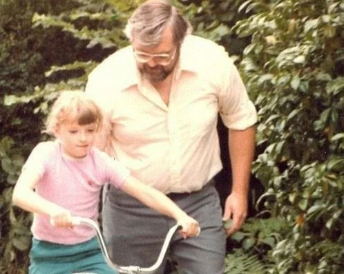 Image of a Dad in white button up shirt teaching young daughter with pink shirt to ride her bike