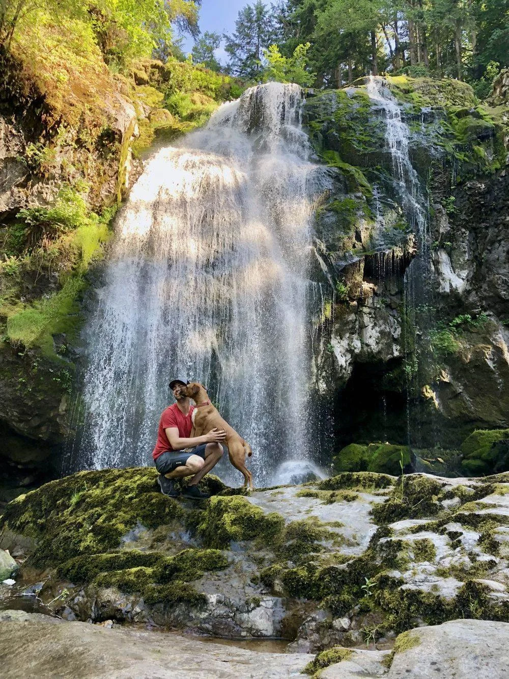 Trey Foster crouching on a mossy rock beside a waterfall while a brown dog leans up to lick his face